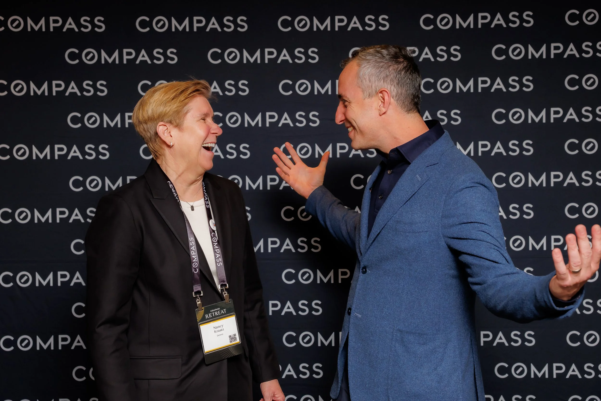 Two professionals, a woman and a man, are smiling and engaging in a conversation at a conference with a backdrop that says 'COMPASS'. The woman is wearing a black blazer, white top, and lanyard, while the man is dressed in a blue blazer and dark shirt.