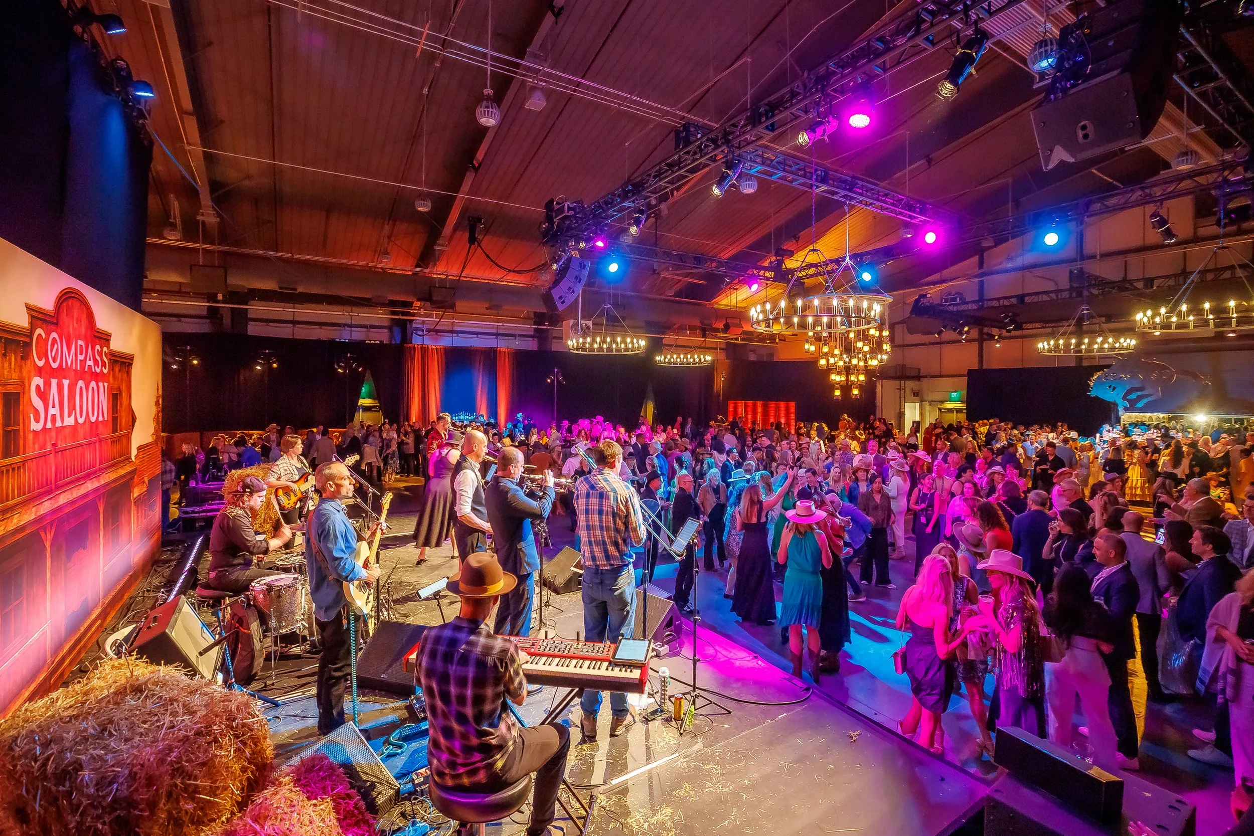 Country-themed dance event with a stage, live band playing instruments, and many people dancing and socializing in a large hall decorated with chandeliers and warm lighting.
