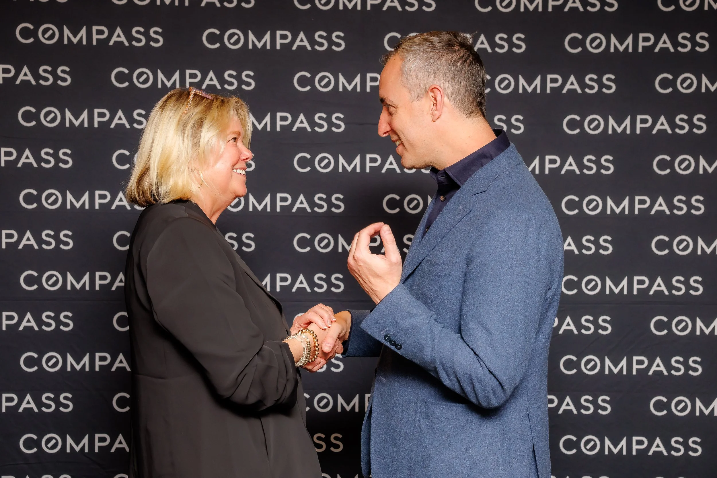 A man and woman smiling and shaking hands in front of a black backdrop with the word 'COMPASS' repeated in white text, while looking at each other.