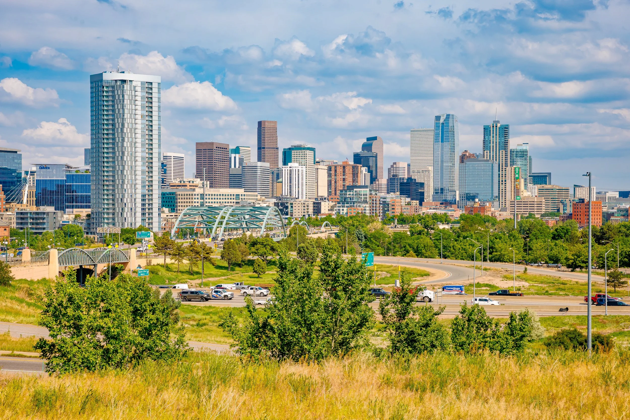 City skyline with tall buildings, green trees, and a highway with cars, under a partly cloudy sky.