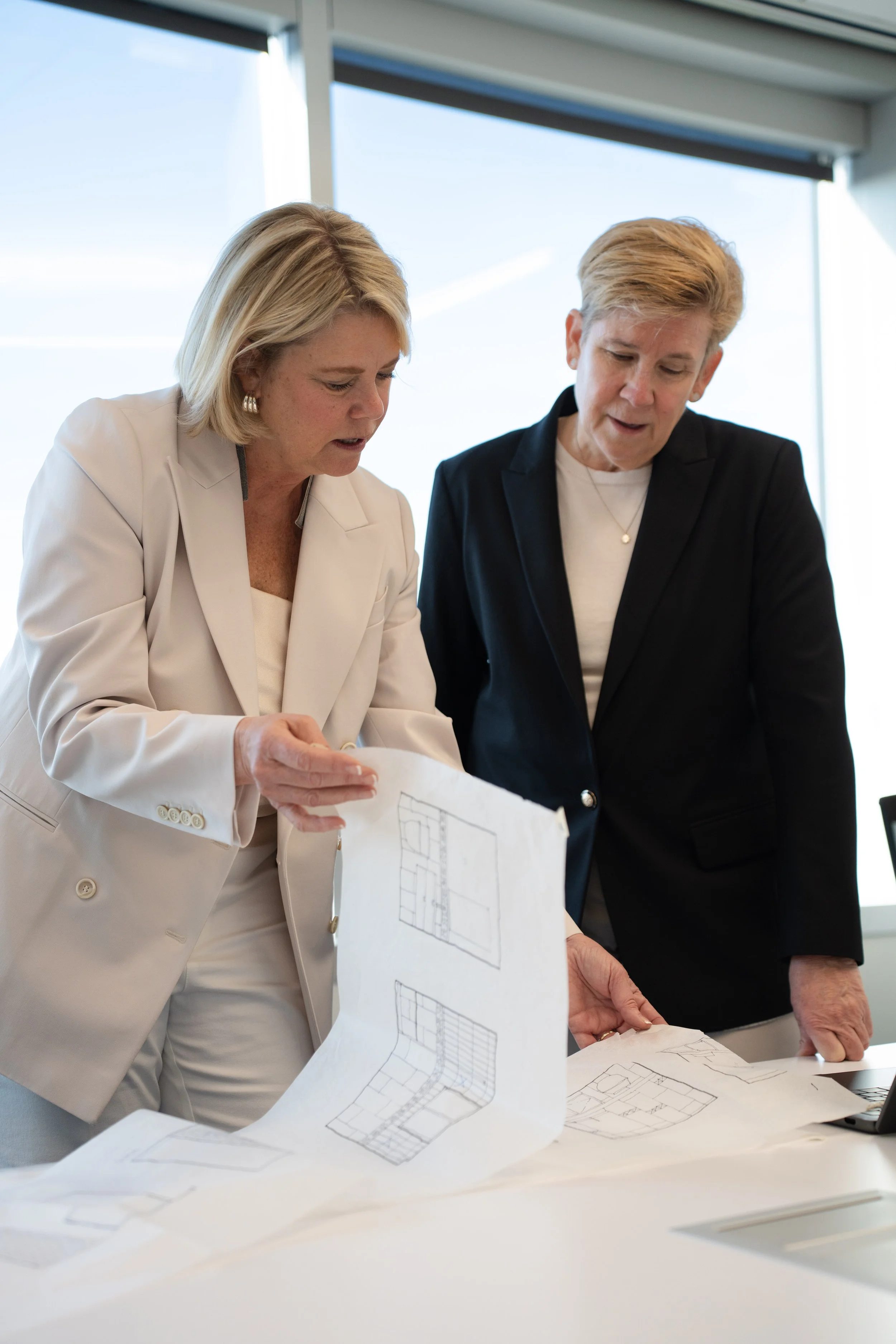 Two women looking at architectural blueprints on a table in a bright office.