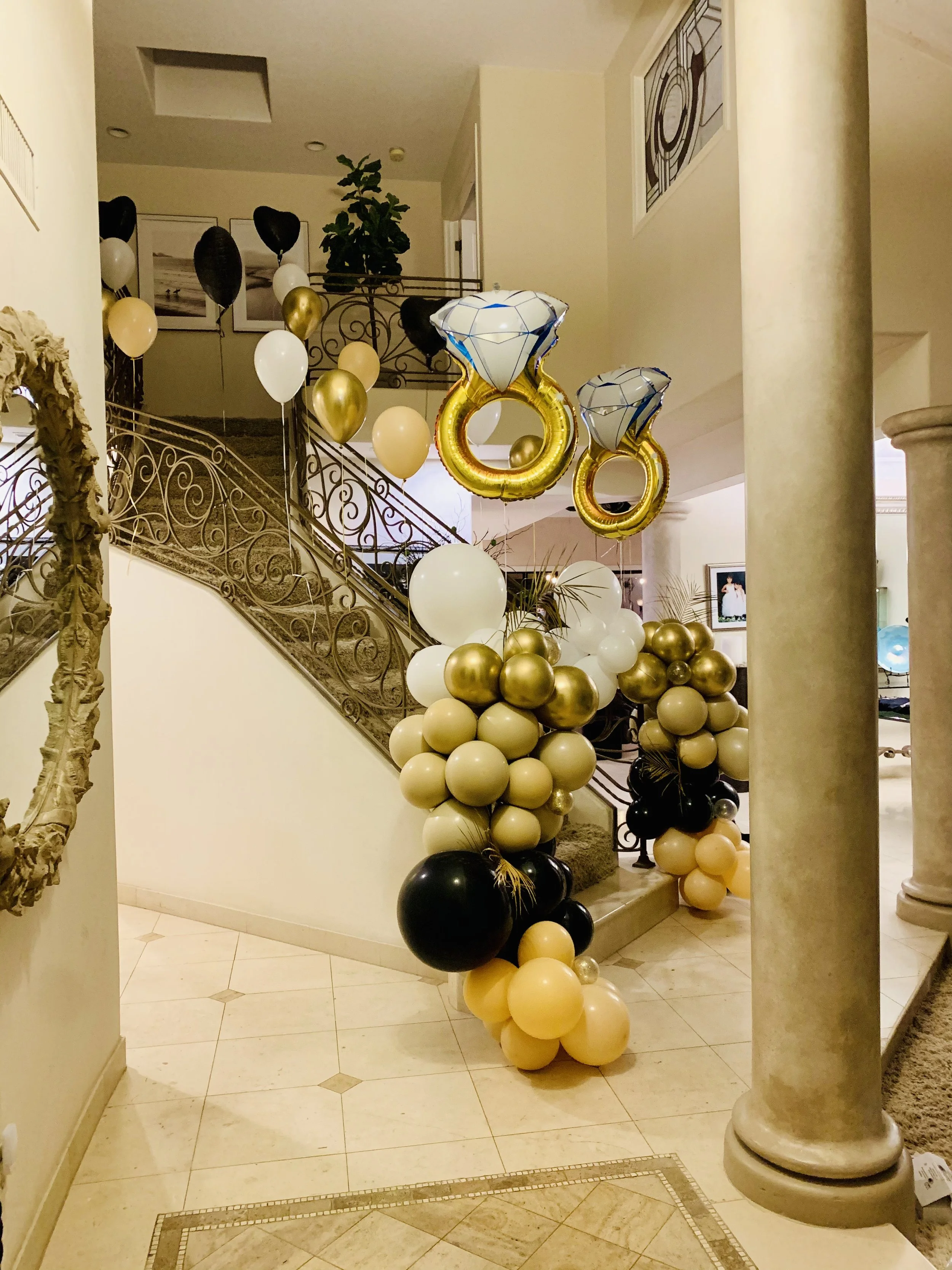 Decorative balloon arrangement with black, gold, cream, and white balloons, including large balloon shaped like a diamond ring, displayed near a staircase with ornate railing, inside a warmly lit room with beige walls, columns, and framed artwork.