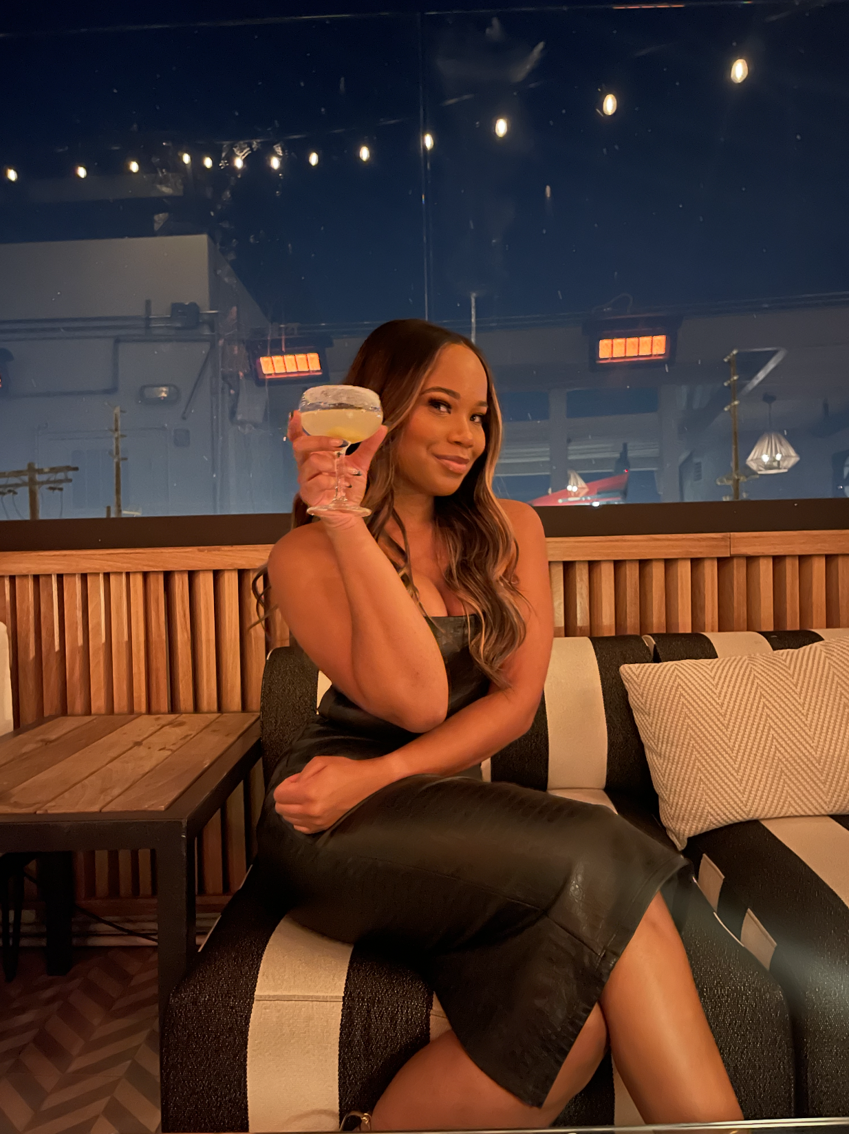A woman with long wavy hair sitting on a black and white striped sofa in a dimly lit room, holding a margarita glass with a salted rim and lemon slice, smiling at the camera.
