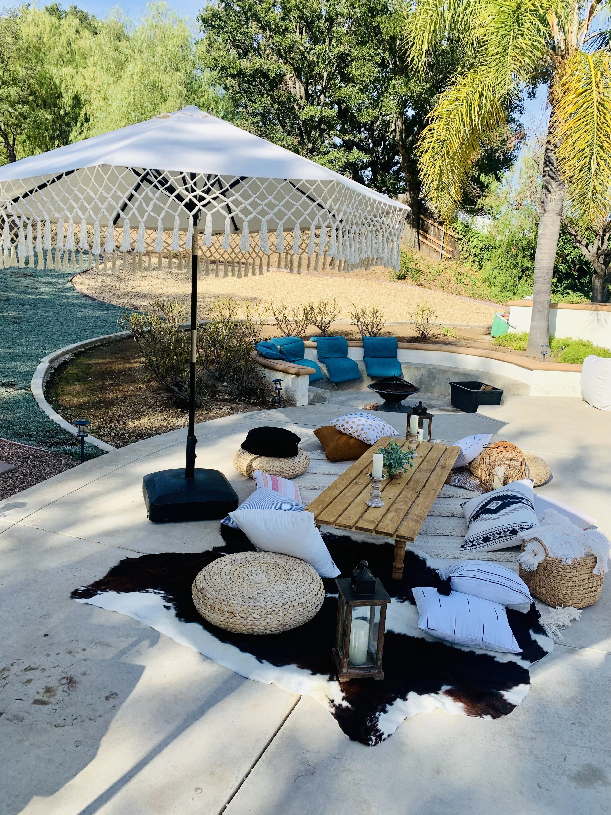 Outdoor patio setup with a large white umbrella, blue cushioned seating, pillows, a wooden table, candles, lanterns, and decorative pillows on a black and white animal hide rug. Surrounding trees and bushes provide greenery.