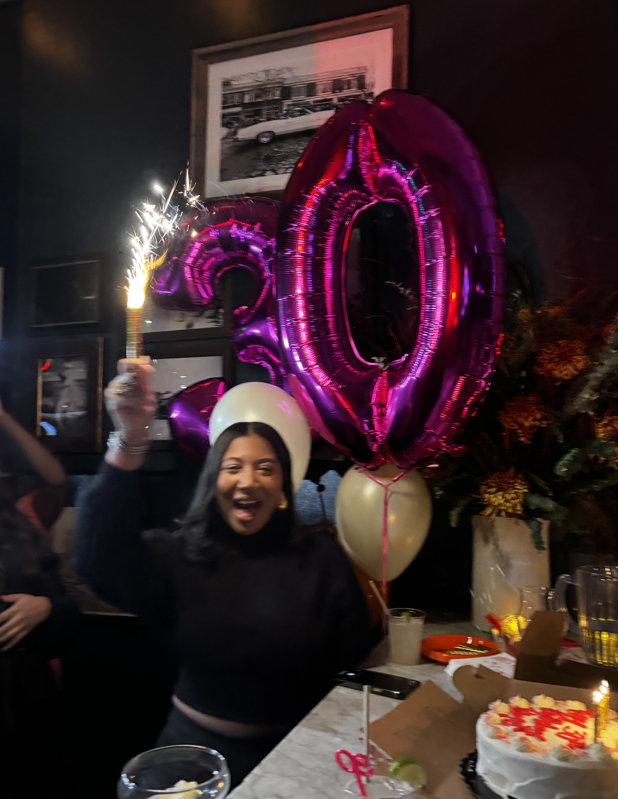 A woman celebrating at a party with balloons, including a large pink number '20', a cake with red and white decorations, and a sparkler in her hand. She is smiling and wearing a white balloon hat.