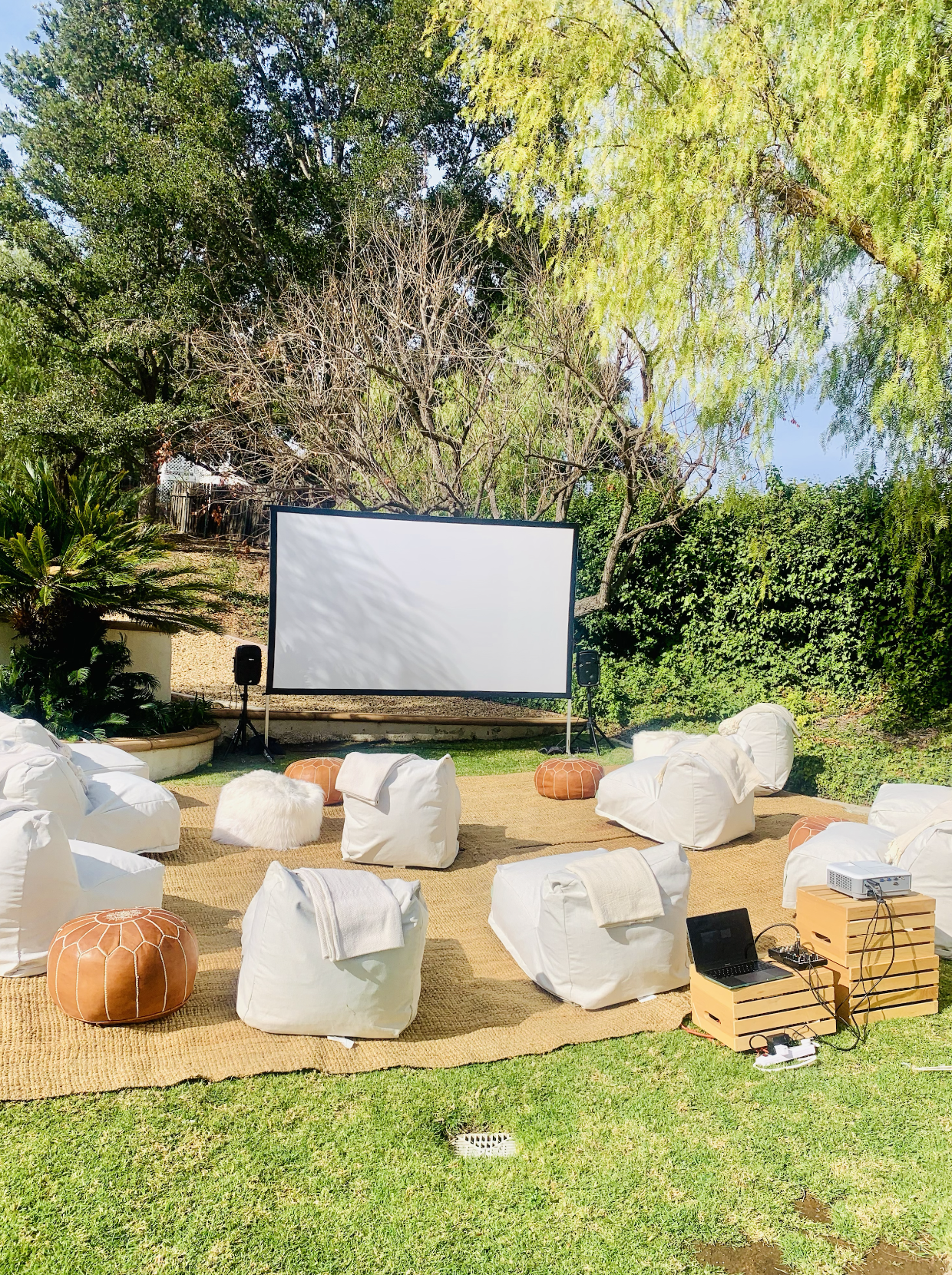 Outdoor movie screening setup with a large white projection screen, surrounded by bean bag chairs and stools on a grassy area under trees with green leaves.