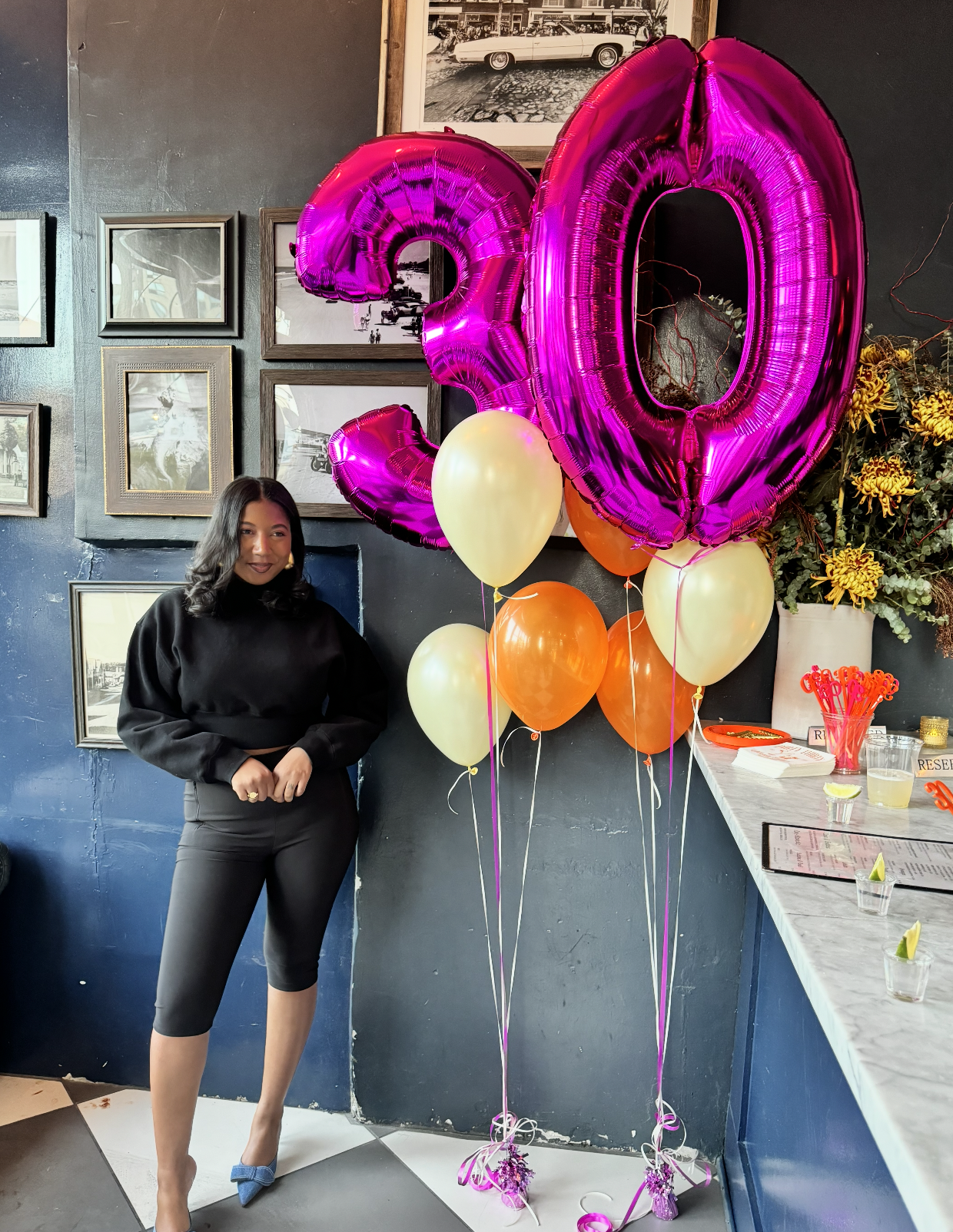 A woman stands near a bouquet of pink, yellow, and orange balloons, with a large pink "20" balloon in the background. She has shoulder-length black hair, wears a black top, black cropped pants, and blue shoes. The setting appears to be a decorated pa