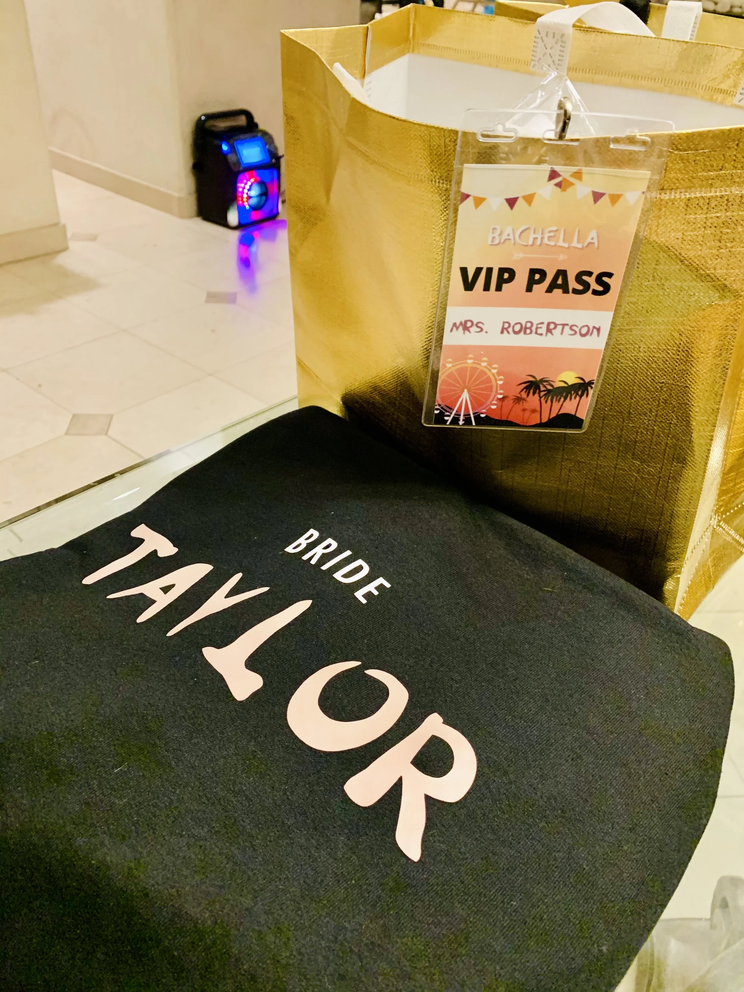 A black tote bag with white text reading 'TRAVEL' is placed on a dark surface. Behind it, there is a gold-colored gift bag with a VIP pass hanging from it. The VIP pass reads 'BACHELLA VIP PASS MRS. ROBERTSON' and has a festive design with a Ferris w