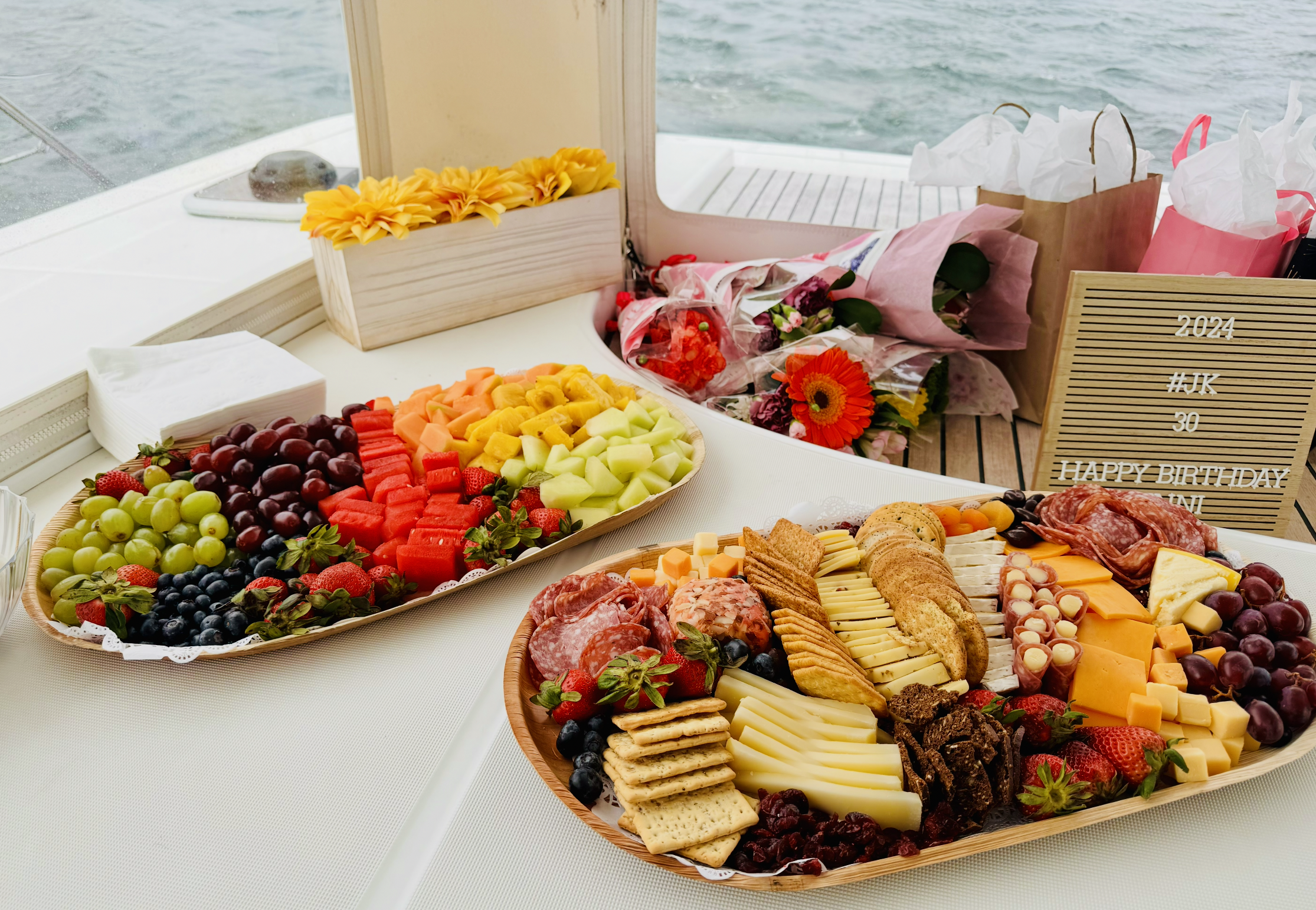 A table of assorted fresh fruits, cheese, crackers, and charcuterie, with a backdrop of water through a window and a birthday sign that reads '2024 #JK 30 HAPPY BIRTHDAY'.