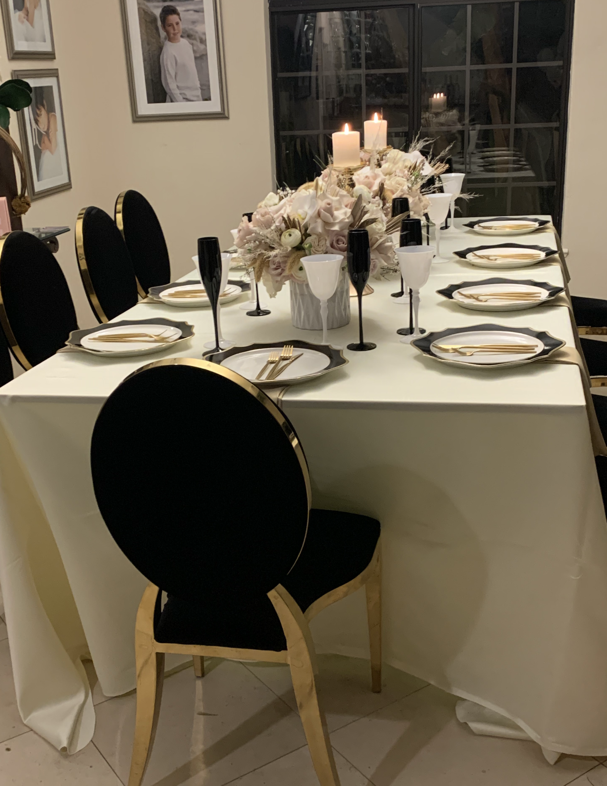 A dining table set for a formal dinner with black and gold chairs, white and black plates, gold cutlery, and black champagne flutes. The table has a large floral centerpiece with candles. The background includes framed photographs and a window.