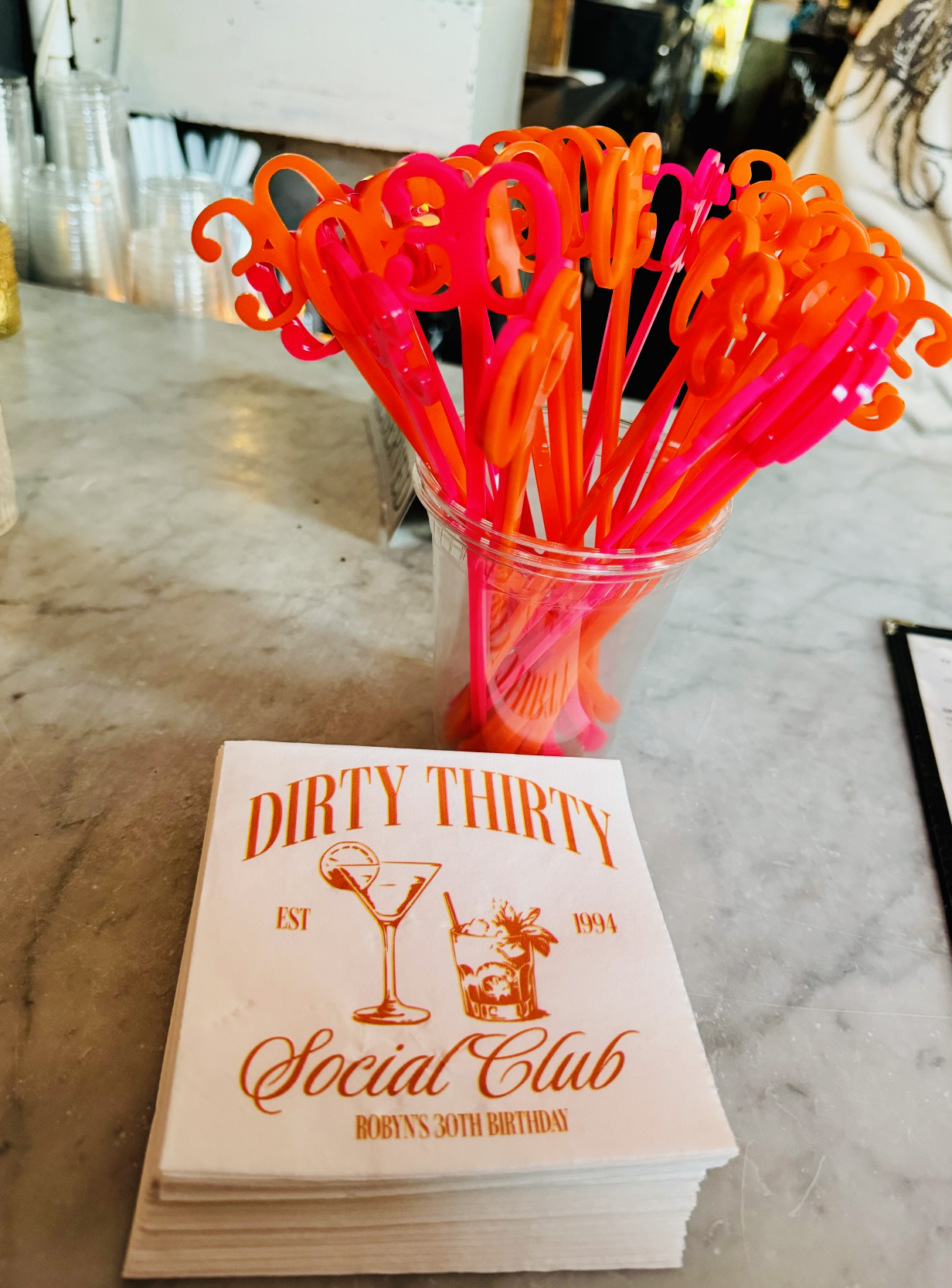 A clear container holding pink and orange curly drink stirrers next to a menu from Dirty Thirty Social Club celebrating Robyn's 30th birthday.