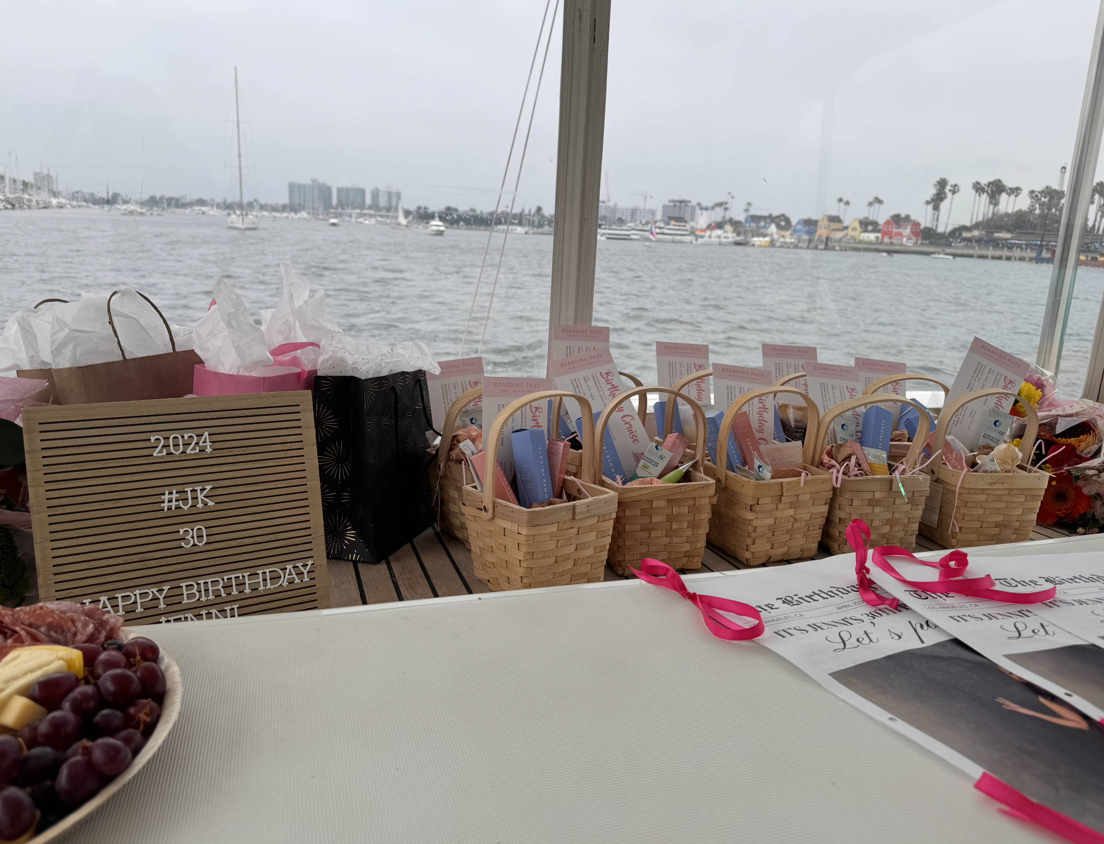 A table decorated for a birthday celebration with baskets of party favors, a sign with the words '2024 Happy Birthday', and a view of a waterfront with boats, buildings, and colorful houses in the background.