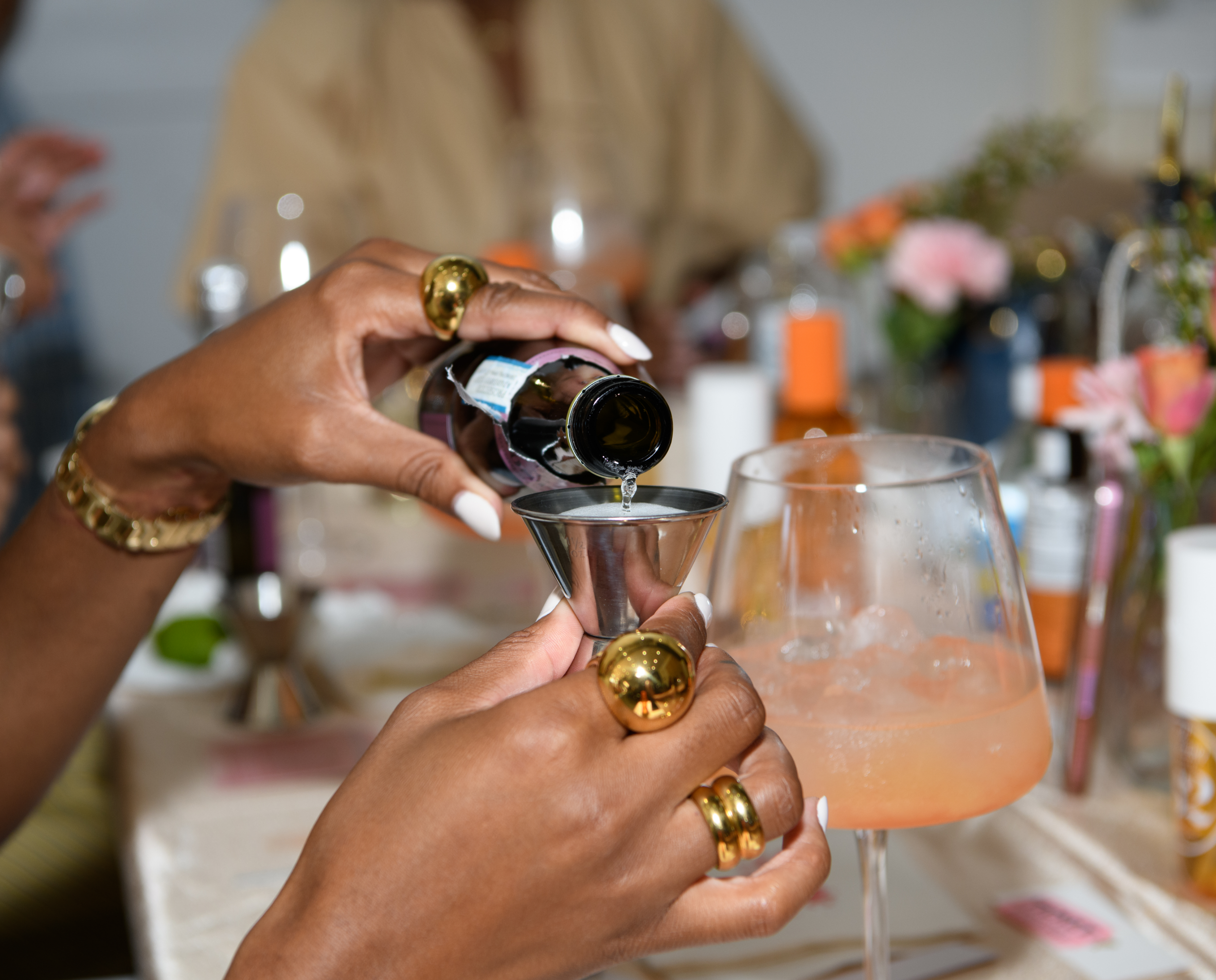 A person with decorated nails and gold jewelry pours a drink into a glass at a celebration or gathering with floral arrangements and various bottles in the background.
