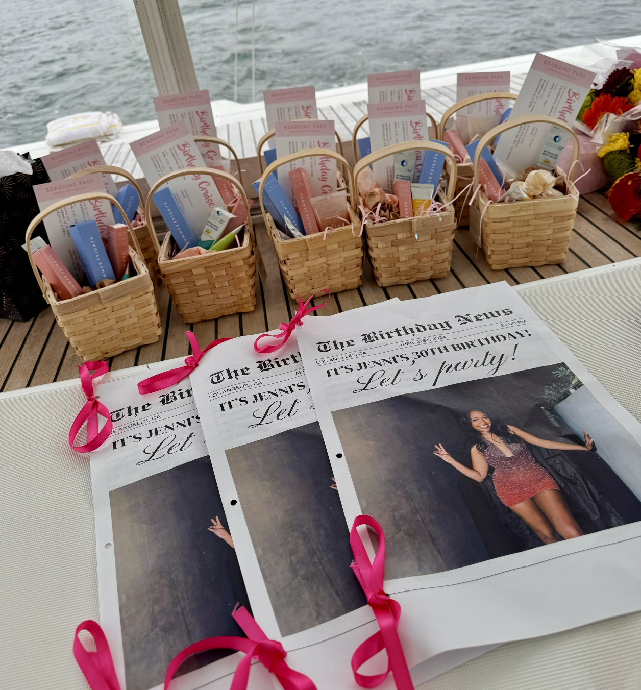 Baskets containing party favors and tickets arranged on a table, with a newspaper announcing a 30th birthday celebration for Jenn, in front of a body of water, possibly on a boat.