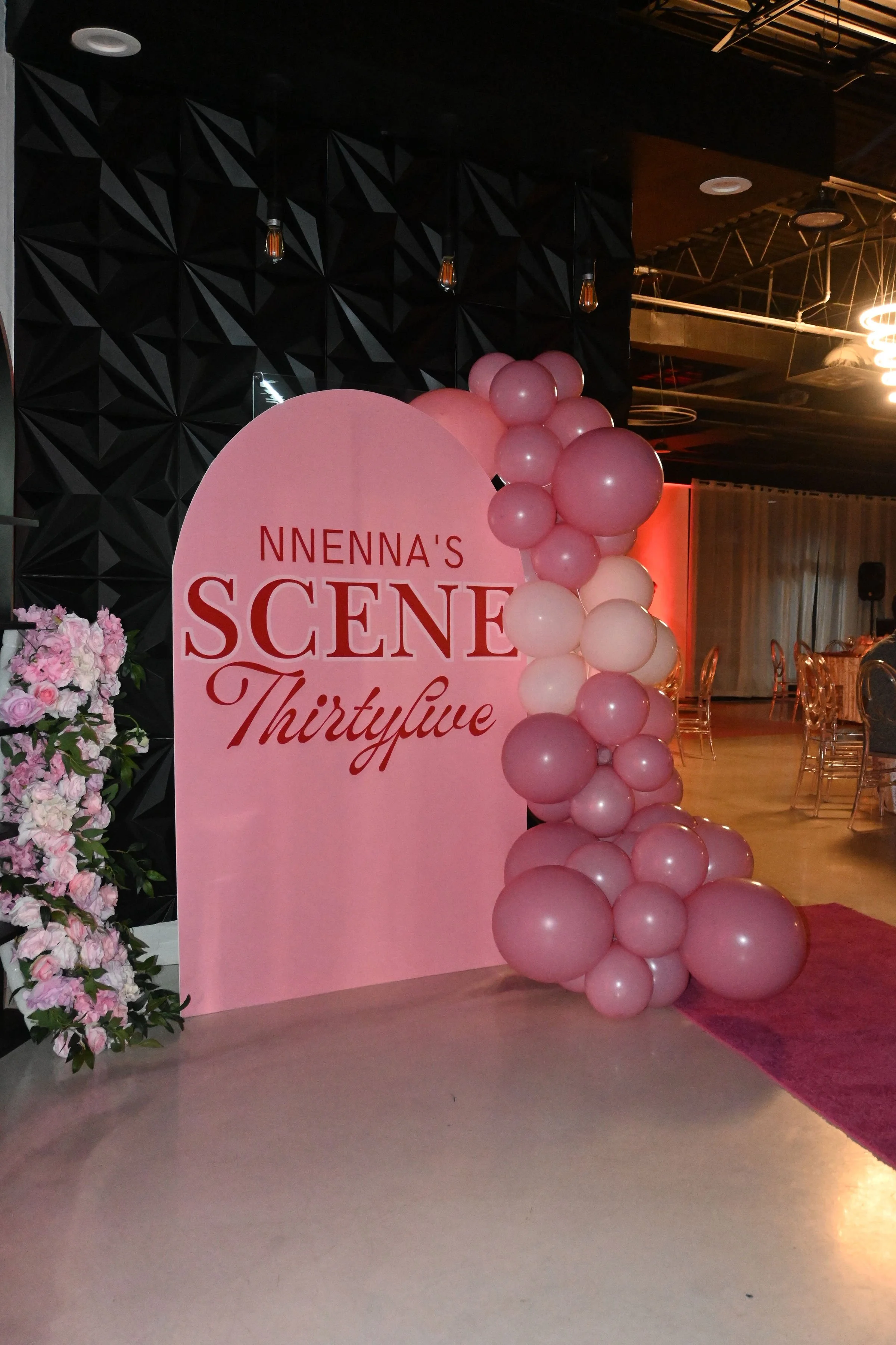 Pink backdrop with balloons on the right side and floral arrangement on the left, at an event celebrating a thirty-fifth birthday.