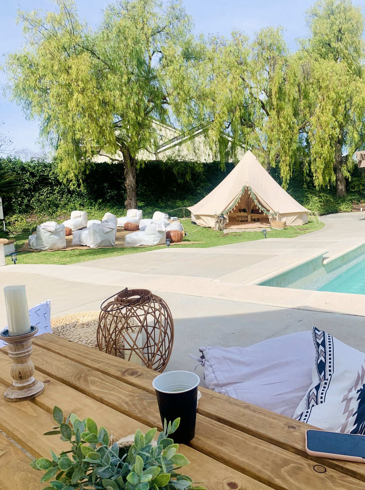Outdoor scene with a swimming pool, a beige tent with decorative garland, white lounge chairs with cushions, green trees, some lanterns, and a wooden table with a candle, a black cup, and a green plant in the foreground.