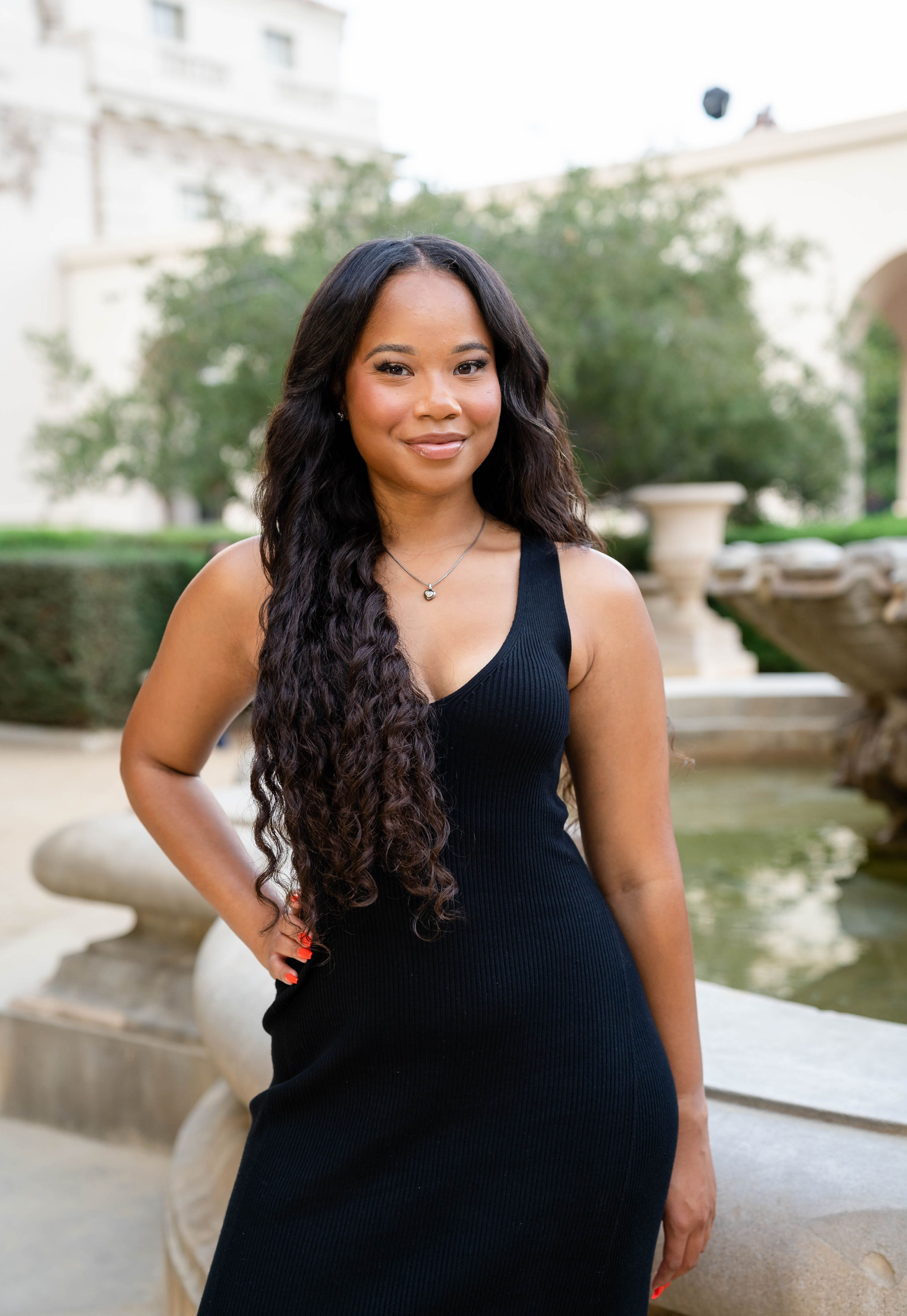 A young woman with long, wavy black hair and a warm smile, wearing a black sleeveless dress and a necklace, stands outdoors in front of a fountain with greenery and white building in the background.