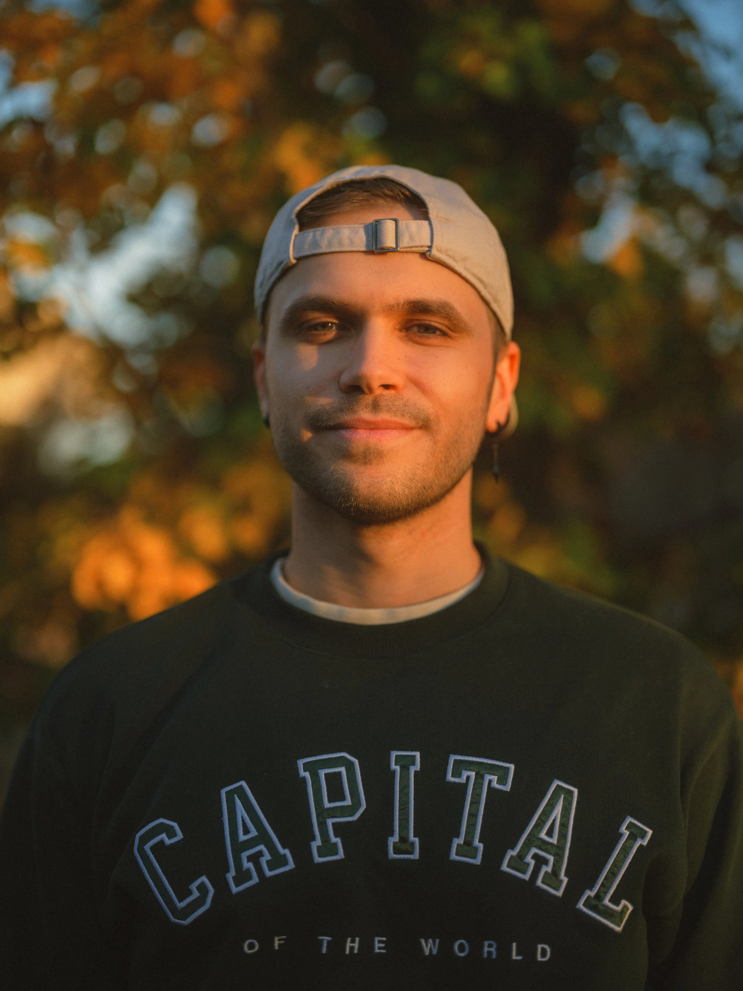 A young man wearing a backward cap and a black sweatshirt with 'CAPITAL' printed on it stands outdoors during sunset, with autumn trees in the background.