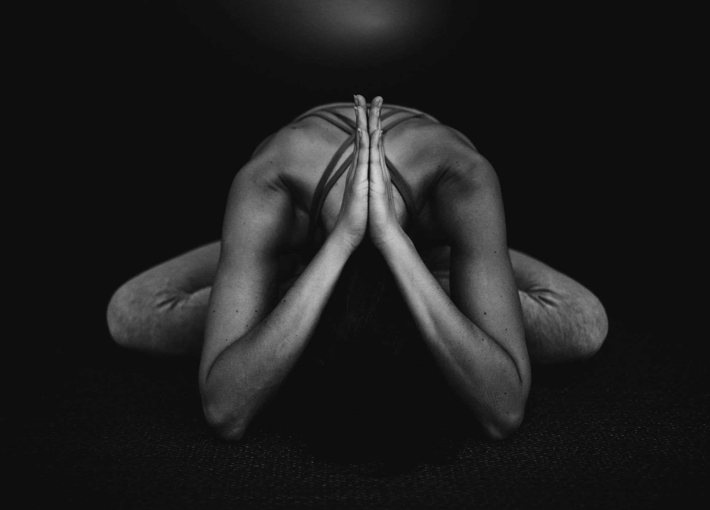 A person practicing yoga in a child's pose, with hands pressed together in front of their face, on a dark surface with a dark background.