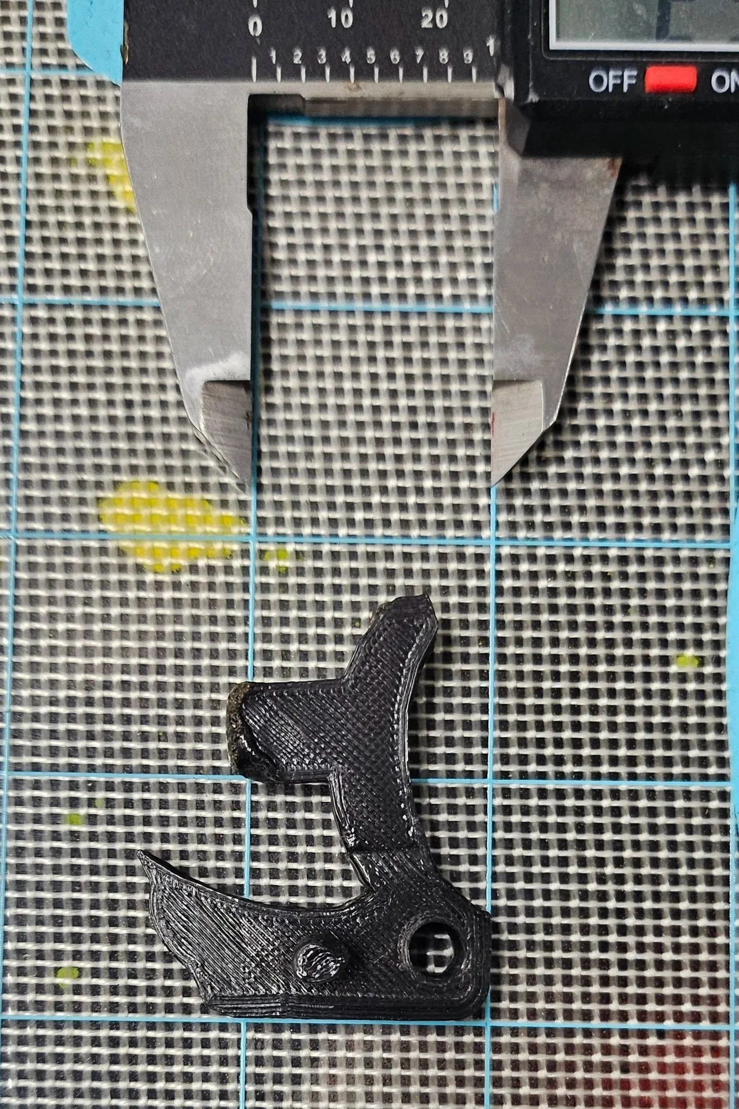 Close-up of a 3D-printed black plastic gun-shaped object placed on a checkered cutting mat, with a metal caliper tool positioned above it.