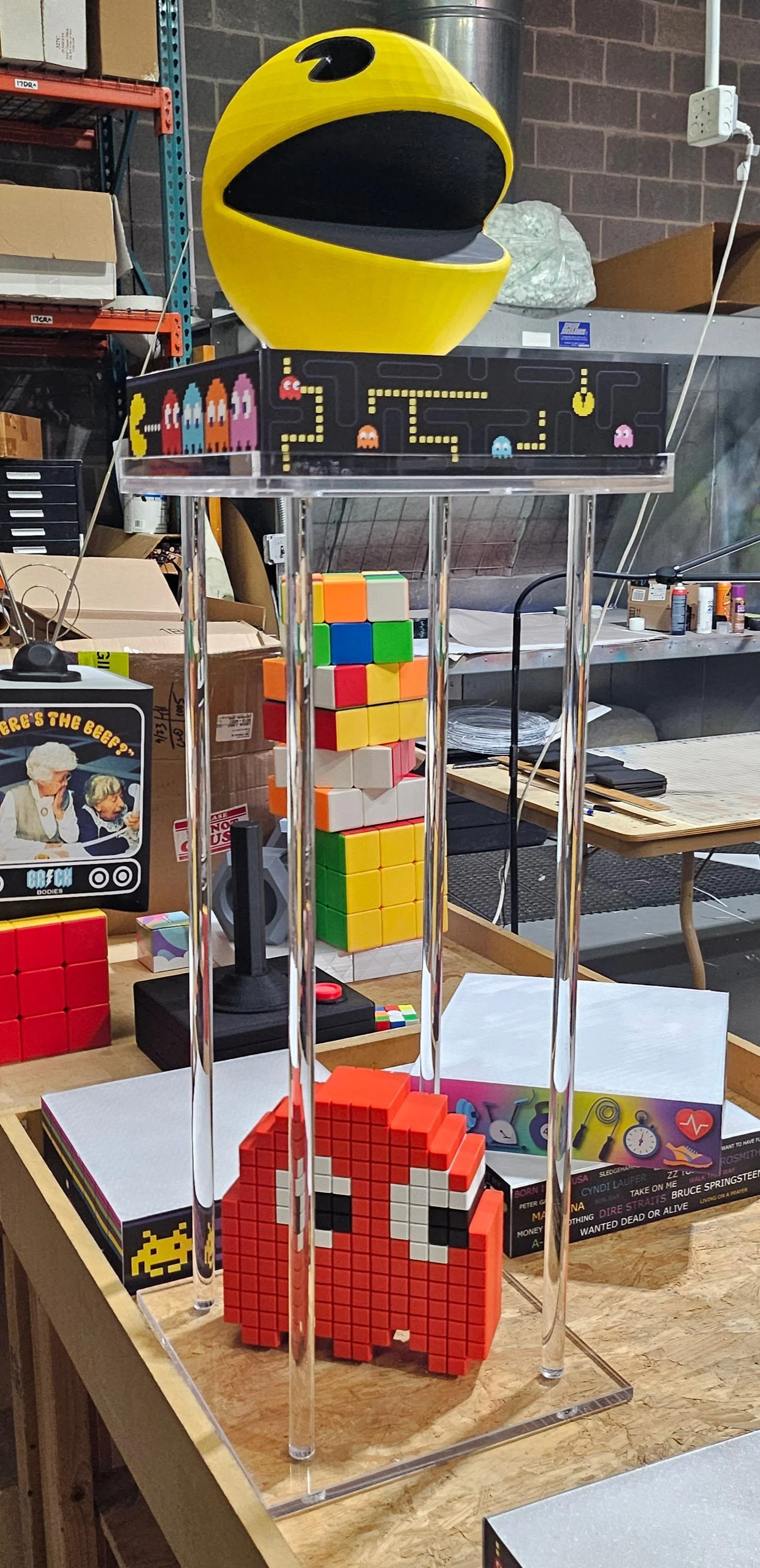 A Pac-Man themed arcade game on top of a clear acrylic stand, with Pac-Man pixel art below and various Rubik's cubes on a worktable, in an industrial workshop setting.
