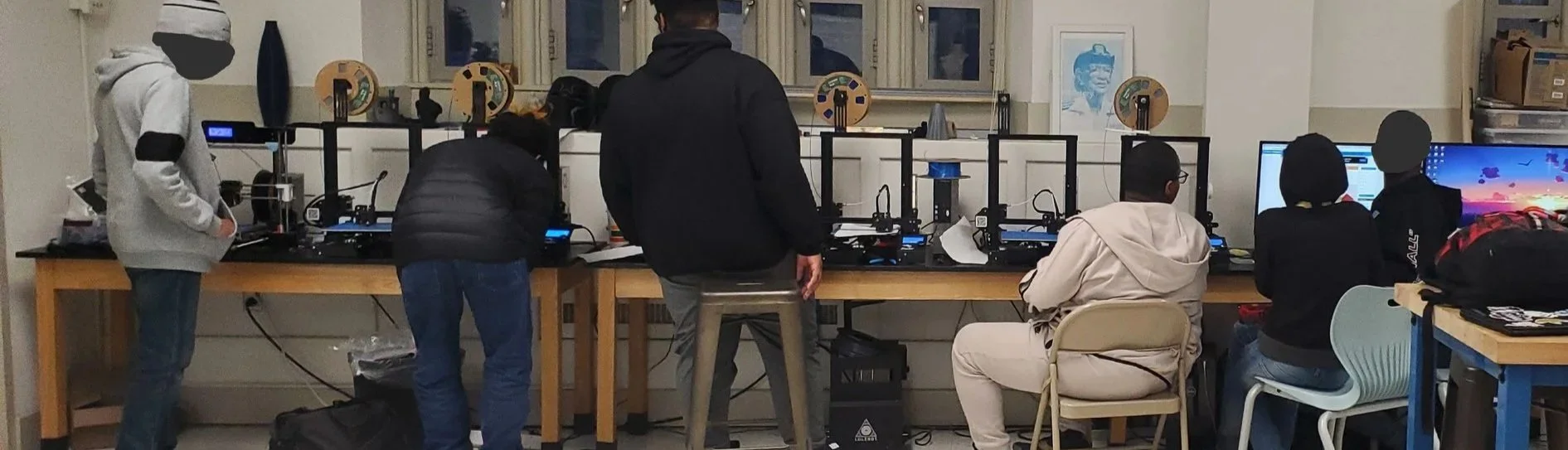 Group of people working with 3D printers in a classroom or workshop setting, some standing and some sitting at desks with computers.