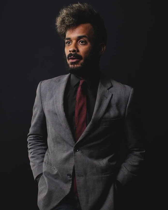 Man in a gray suit, black dress shirt, and maroon tie standing against a dark background.