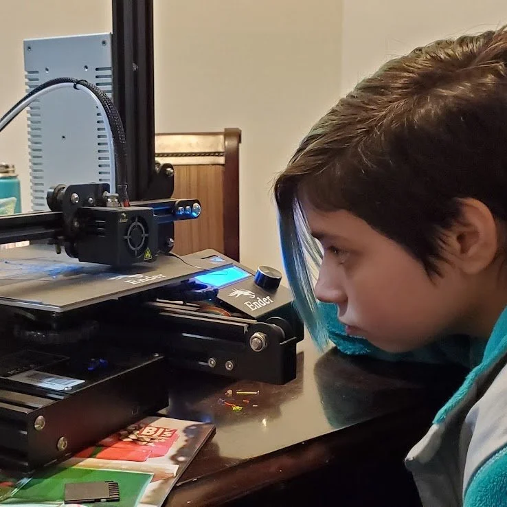 Person using a 3D printer at a desk, focusing intently on the printer with a small spool of filament nearby.