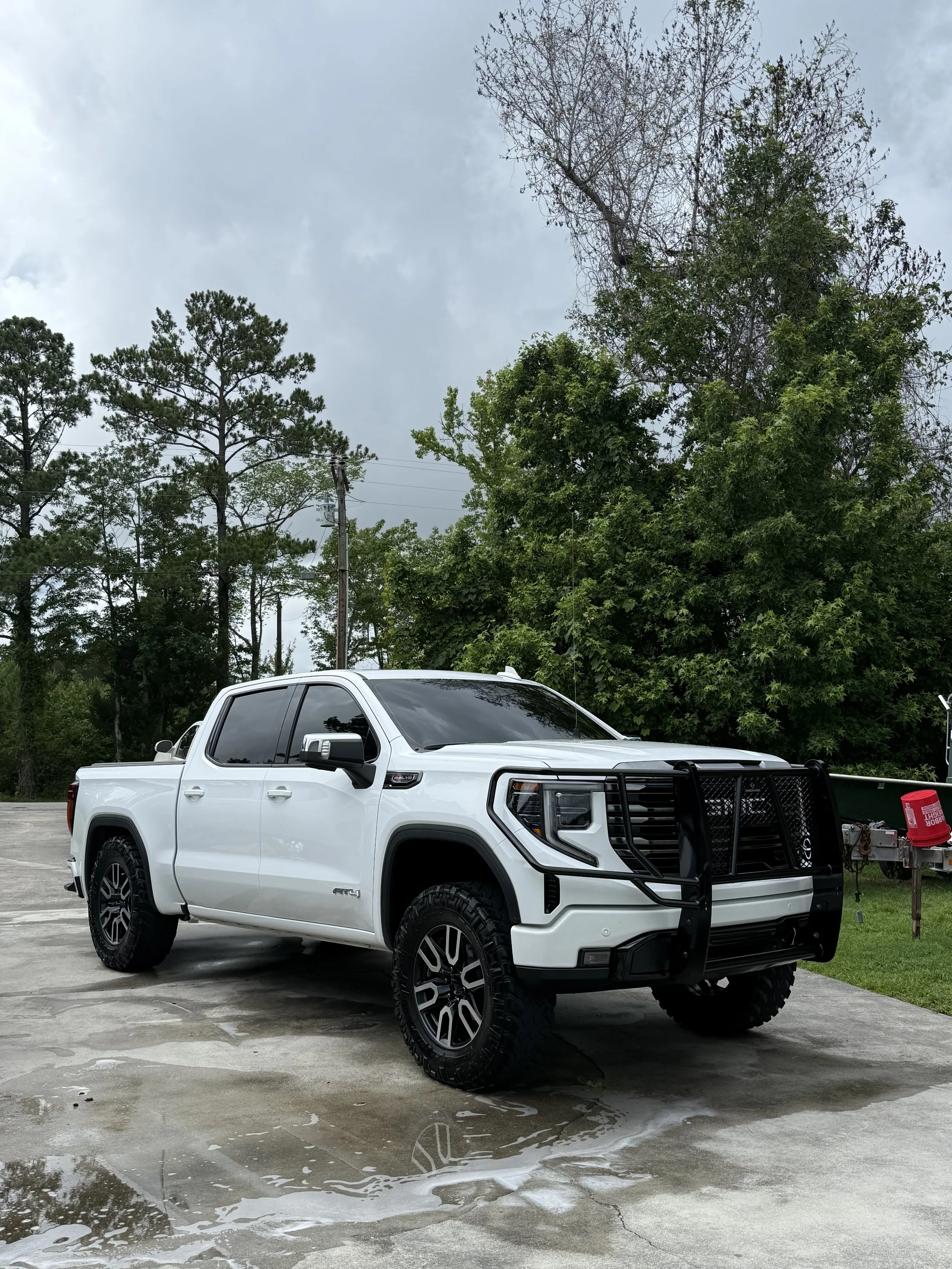 White pickup truck with a black grille guard parked on a wet concrete surface under a cloudy sky, with trees and greenery in the background.