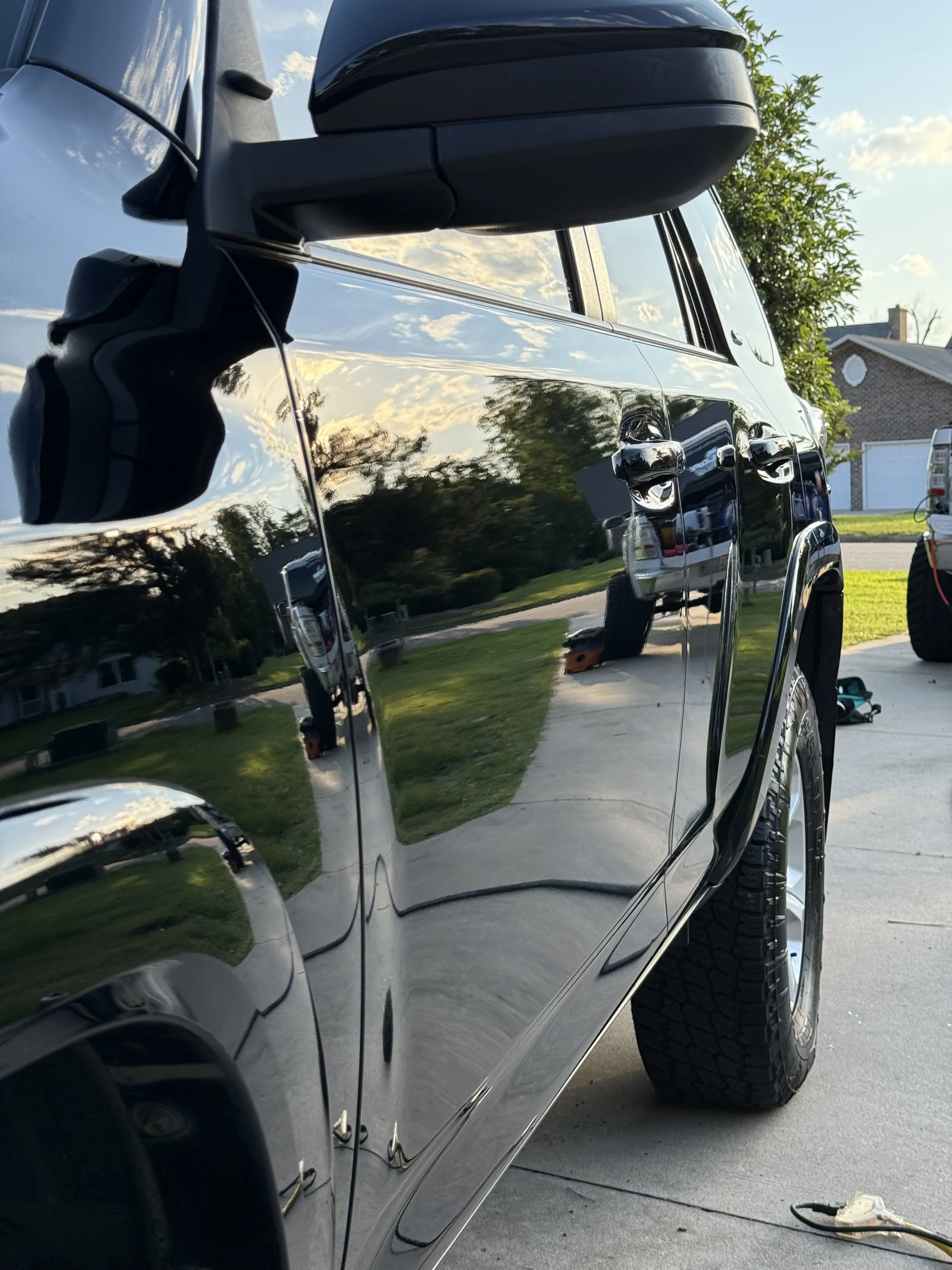 Close-up of a black car's side with a highly reflective surface, showing reflections of the sky, trees, and nearby houses, parked on a driveway in a suburban neighborhood.