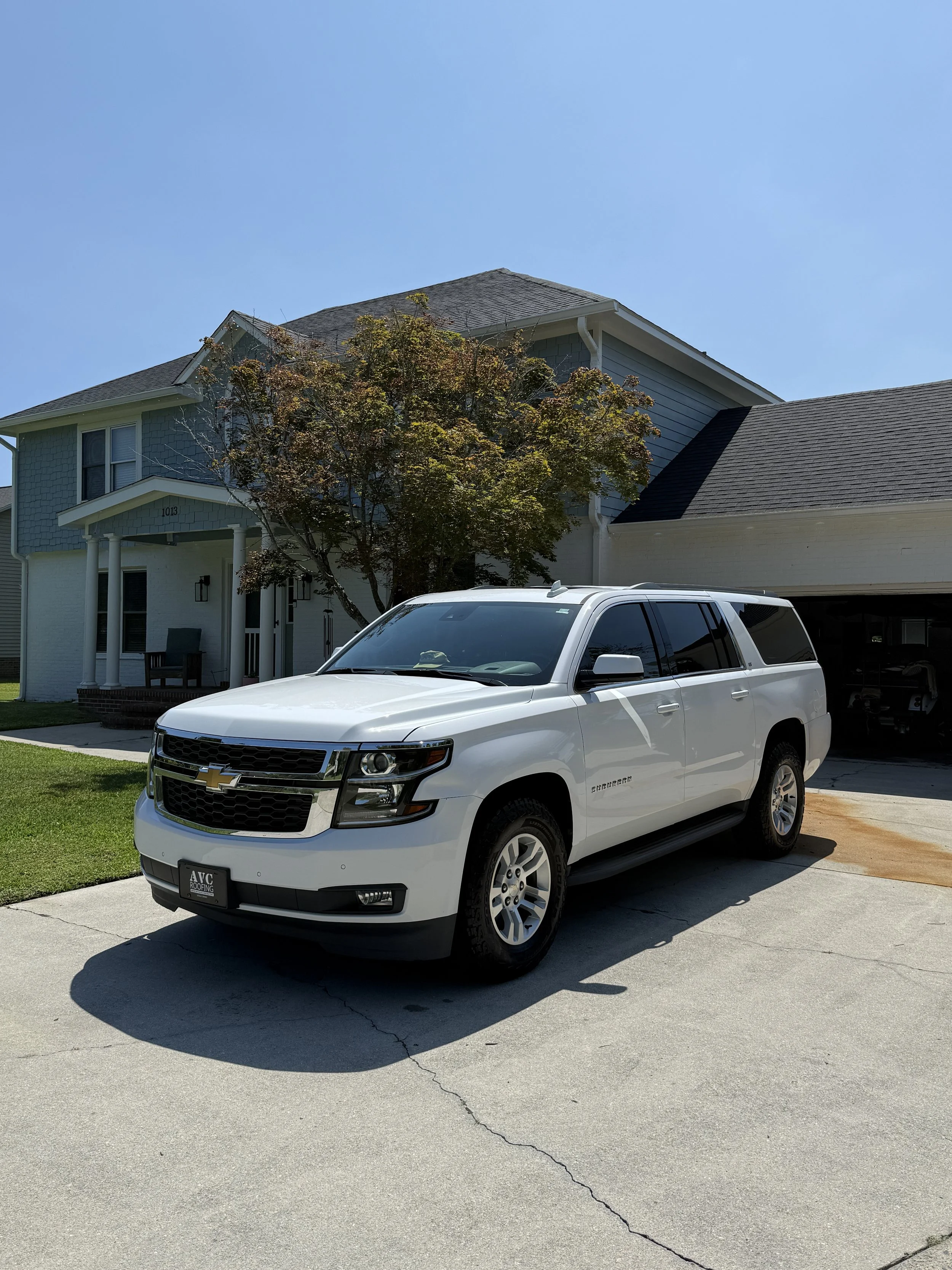 White Chevrolet Suburban parked on driveway in front of a house with a tree and clear blue sky