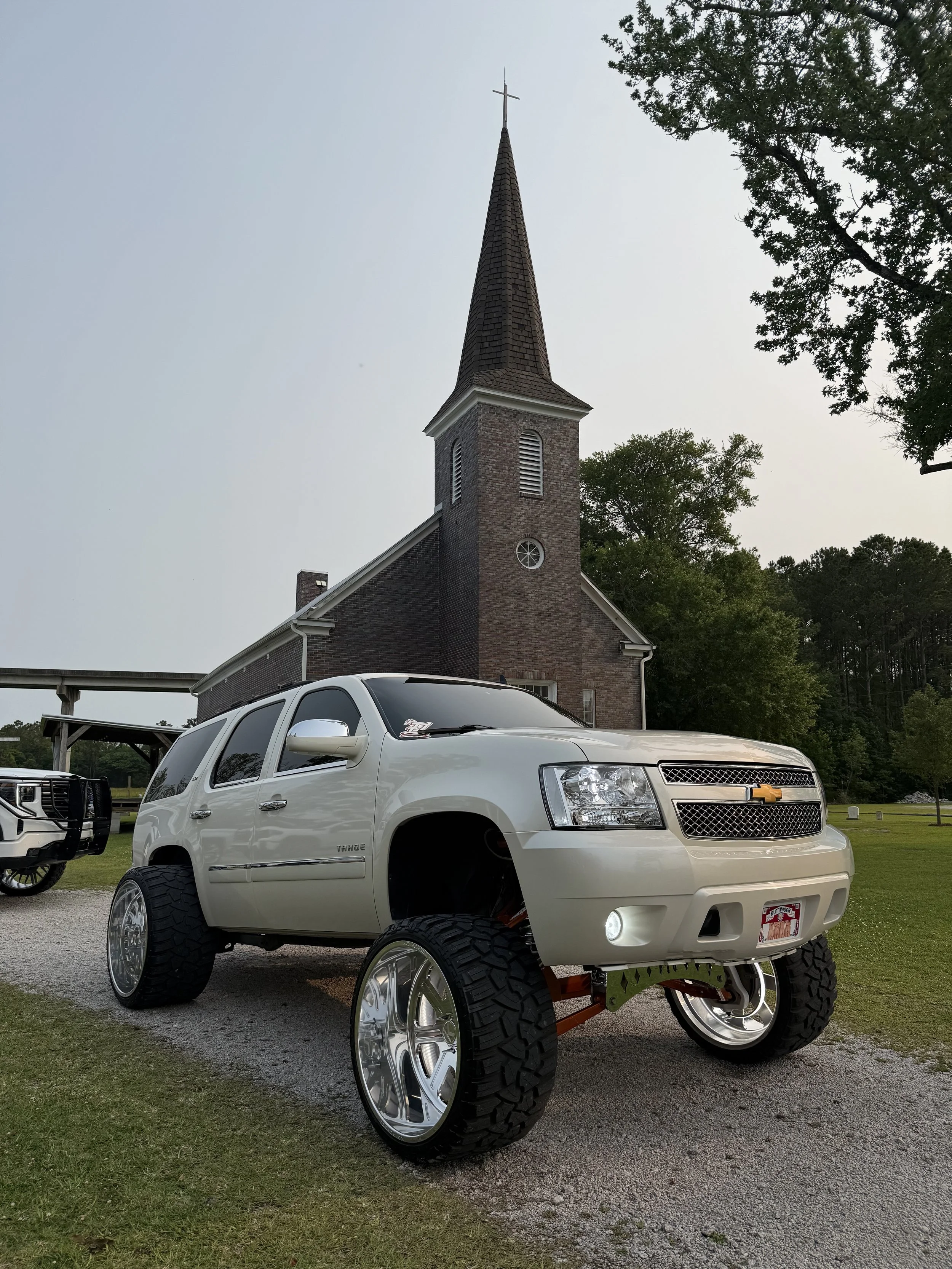 A lifted white Chevrolet Tahoe SUV parked on a gravel lot in front of a brick church with a tall steeple, surrounded by trees.
