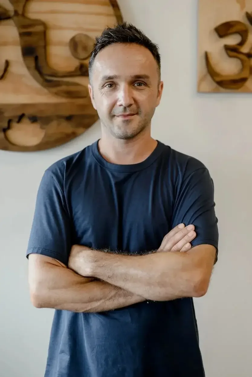 A man with short dark hair and light skin standing indoors with arms crossed, wearing a dark blue t-shirt, smiling slightly, with wooden art and the number 3 in the background.
