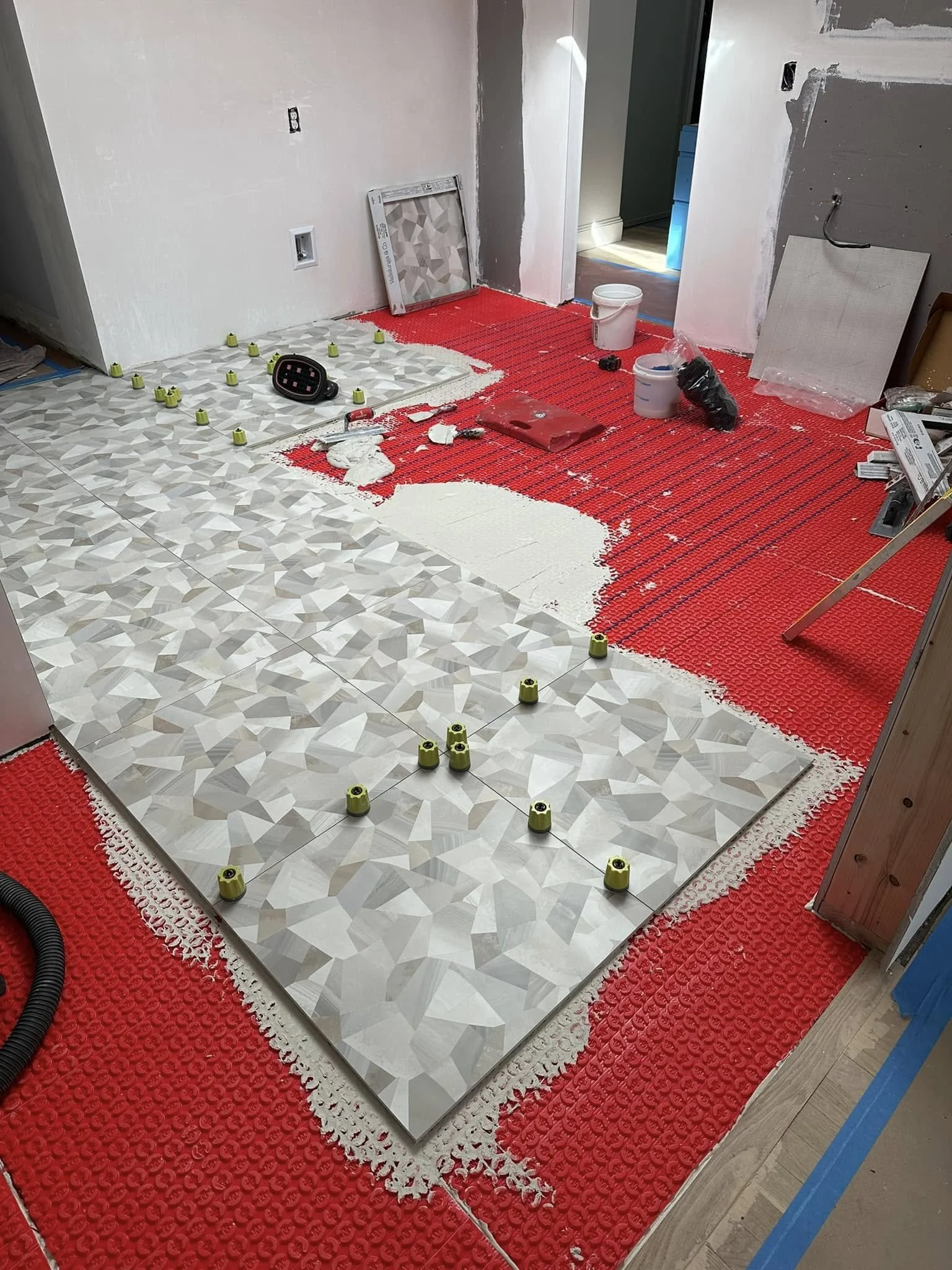 Room under renovation with red uncoupling membrane on the floor, partially installed geometric patterned tiles, and construction tools.