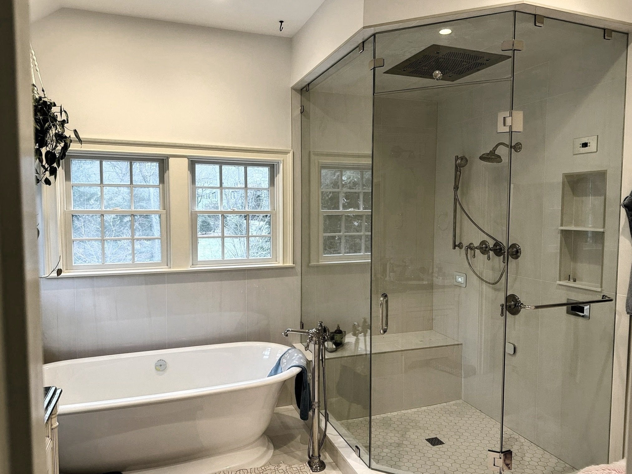 Modern bathroom interior with a freestanding bathtub and glass-enclosed walk-in shower; wall-mounted fixtures and windows are visible.