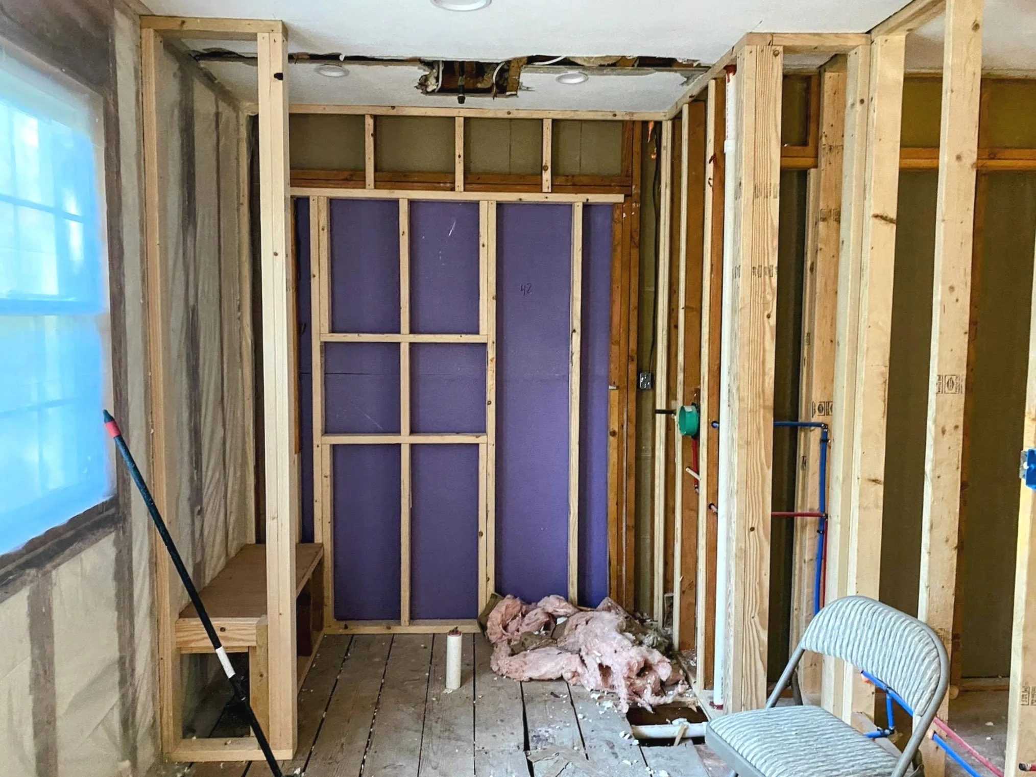 Interior of a room under construction with exposed wooden framing, insulation materials on the floor, purple drywall, and a folding chair.