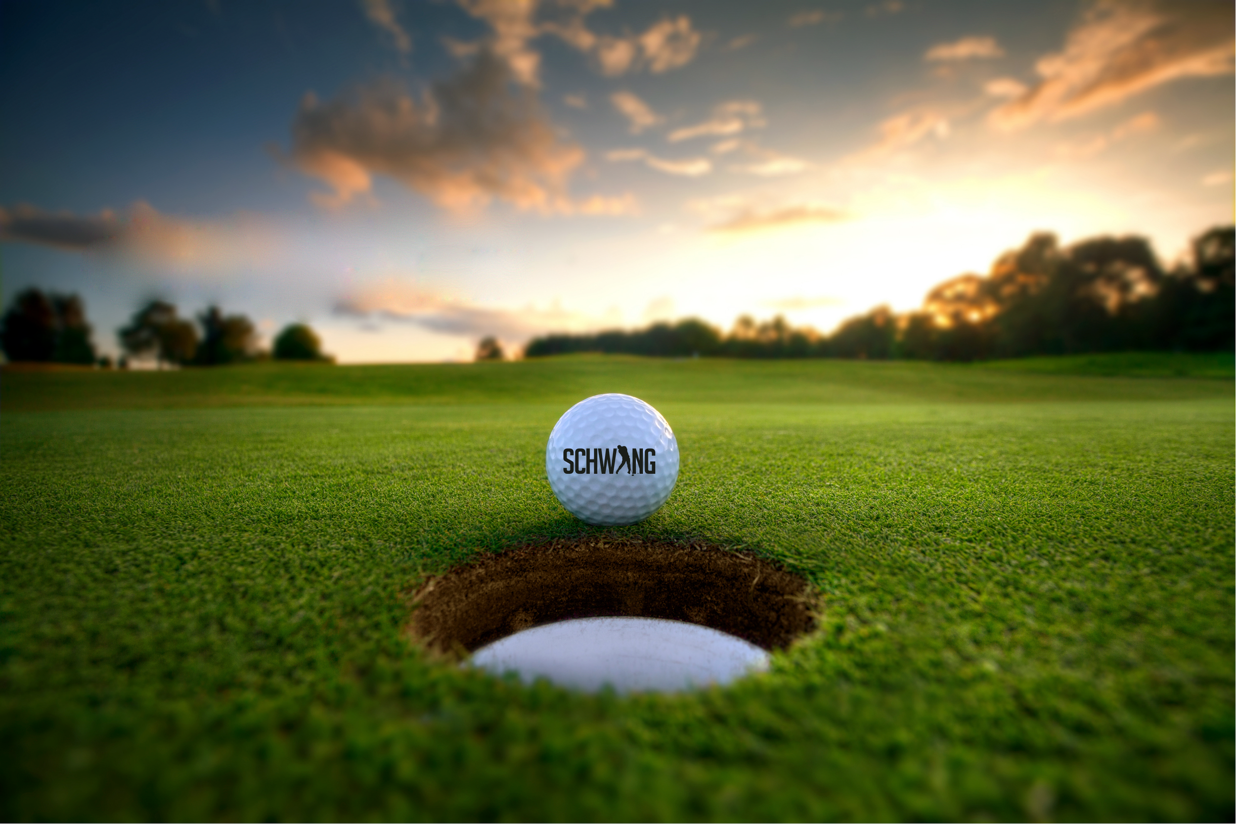 A golf ball with the word 'SCHWING' on it positioned near a hole on a golf course at sunset, with trees in the background and a partly cloudy sky.