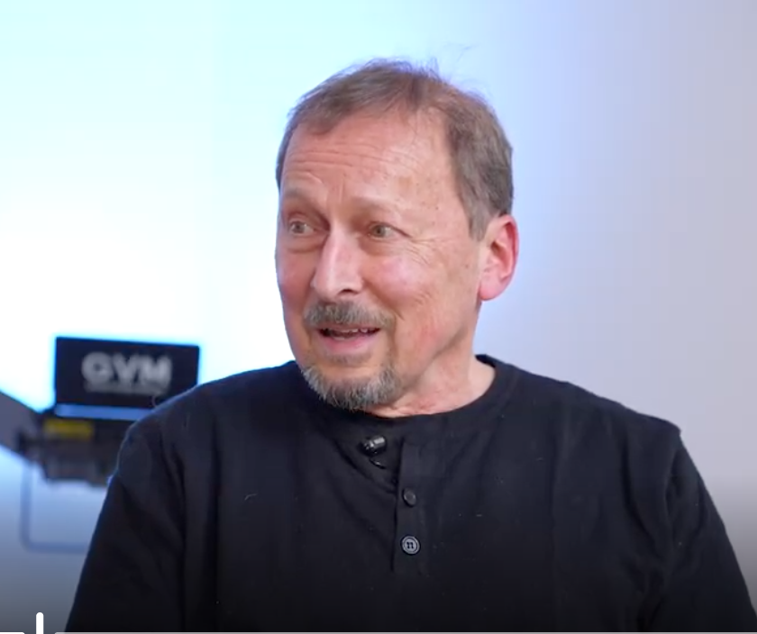 A middle-aged man with light brown hair, a beard, and a mustache, wearing a black shirt, speaking or smiling during an interview in a room with a bright background and a blurry gym sign in the background.