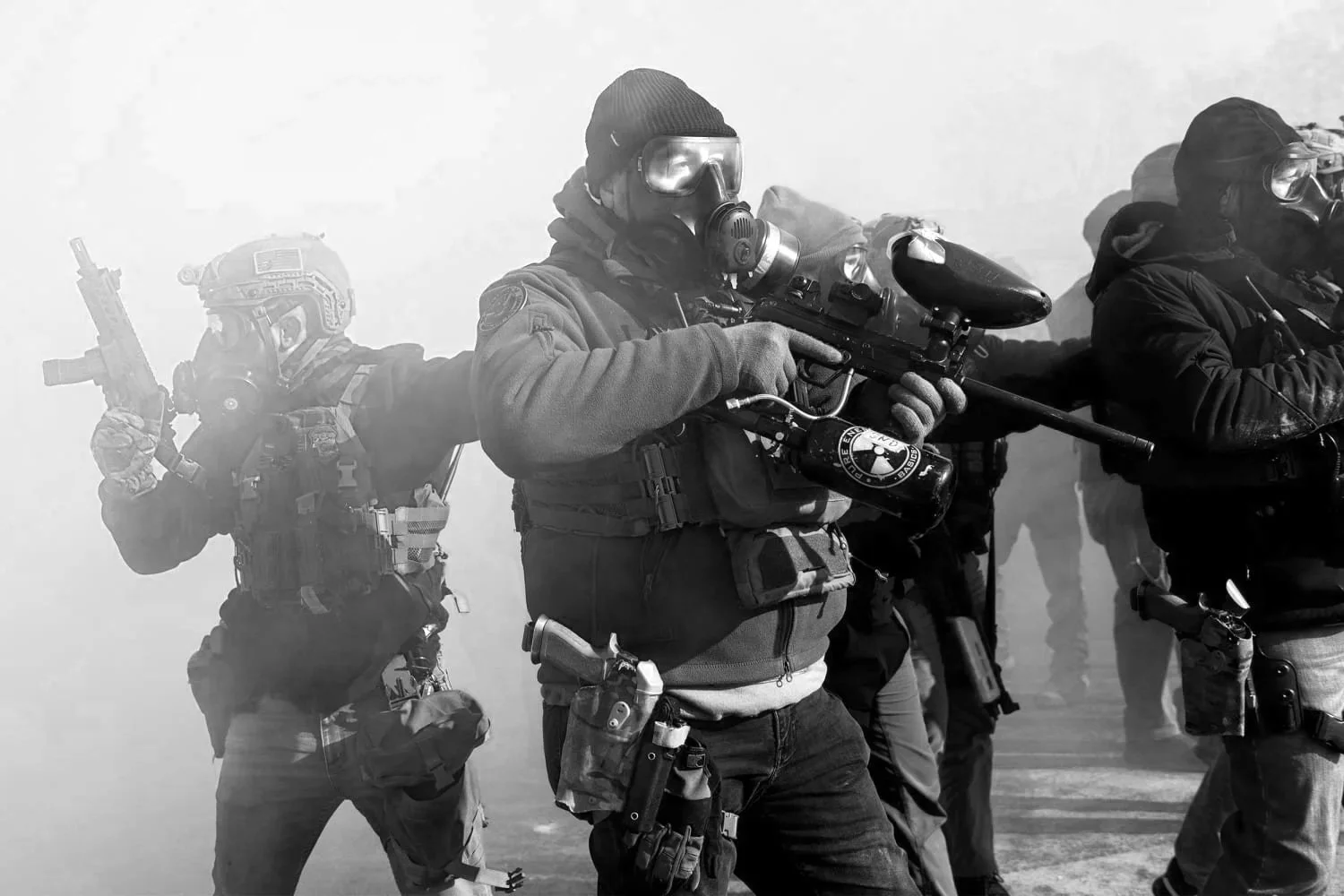 Black and white photo of armed police or military personnel in tactical gear and masks, holding weapons in a tactical formation in a smoky environment.