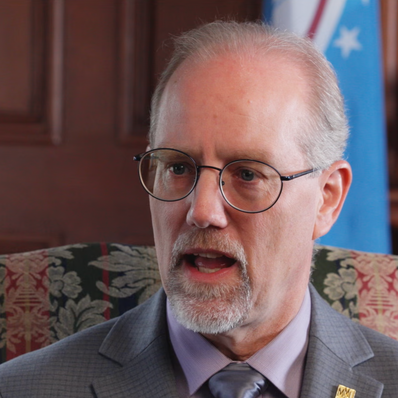 Close-up of a man with gray hair, glasses, and a beard, speaking in an office or formal setting.
