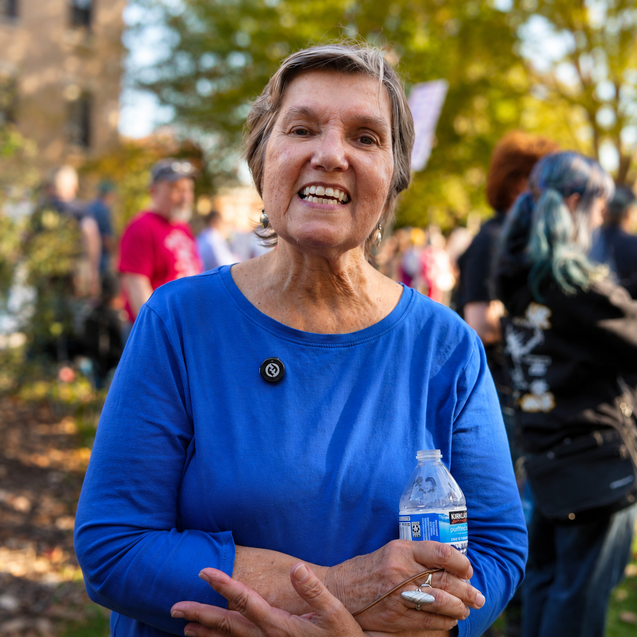 An elderly woman with short gray hair wearing a blue shirt holding a water bottle, standing outdoors among a group of people, with trees and sunlight in the background.