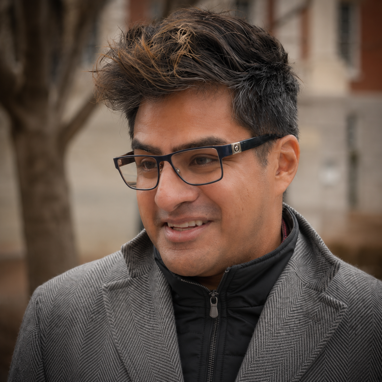 A man with dark hair with lighter streaks, wearing black glasses, a gray blazer, and a black zip-up jacket, smiling outdoors with blurred trees and buildings in the background.