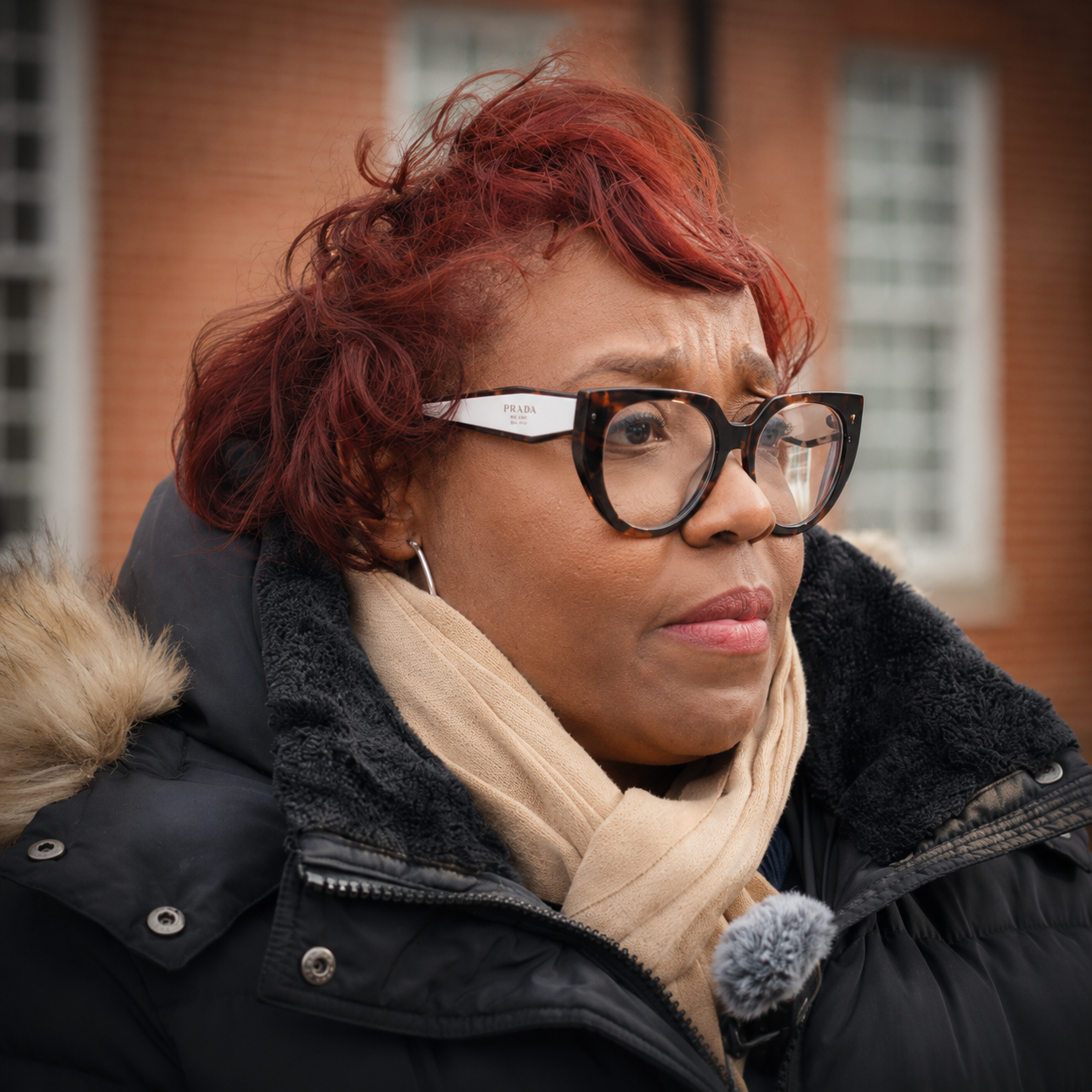 Close-up of a woman with red hair, glasses, and a beige scarf, wearing a black coat with fur trim, outdoors in front of a brick building.