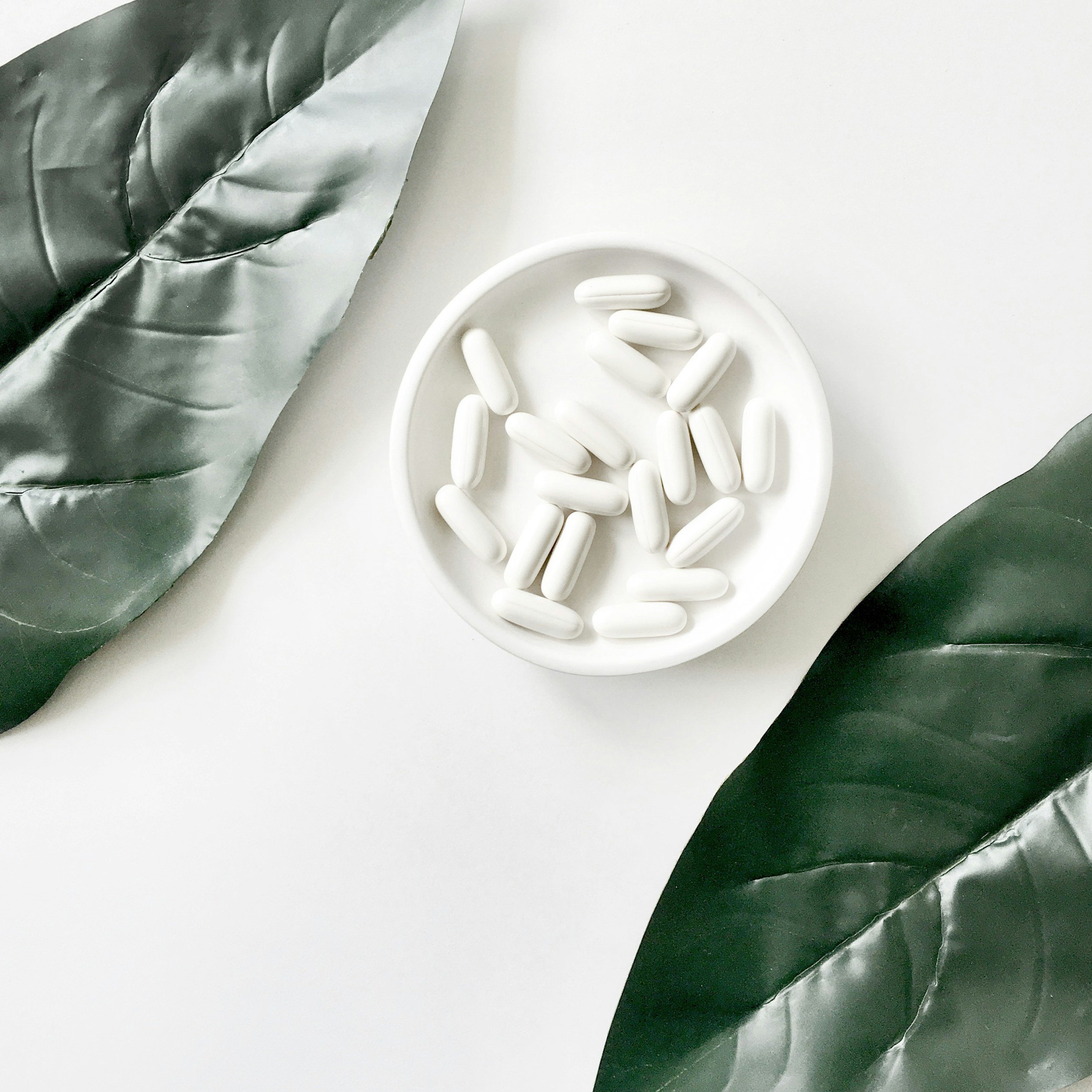 A small white bowl filled with medication tablets, resting on large green leaves.