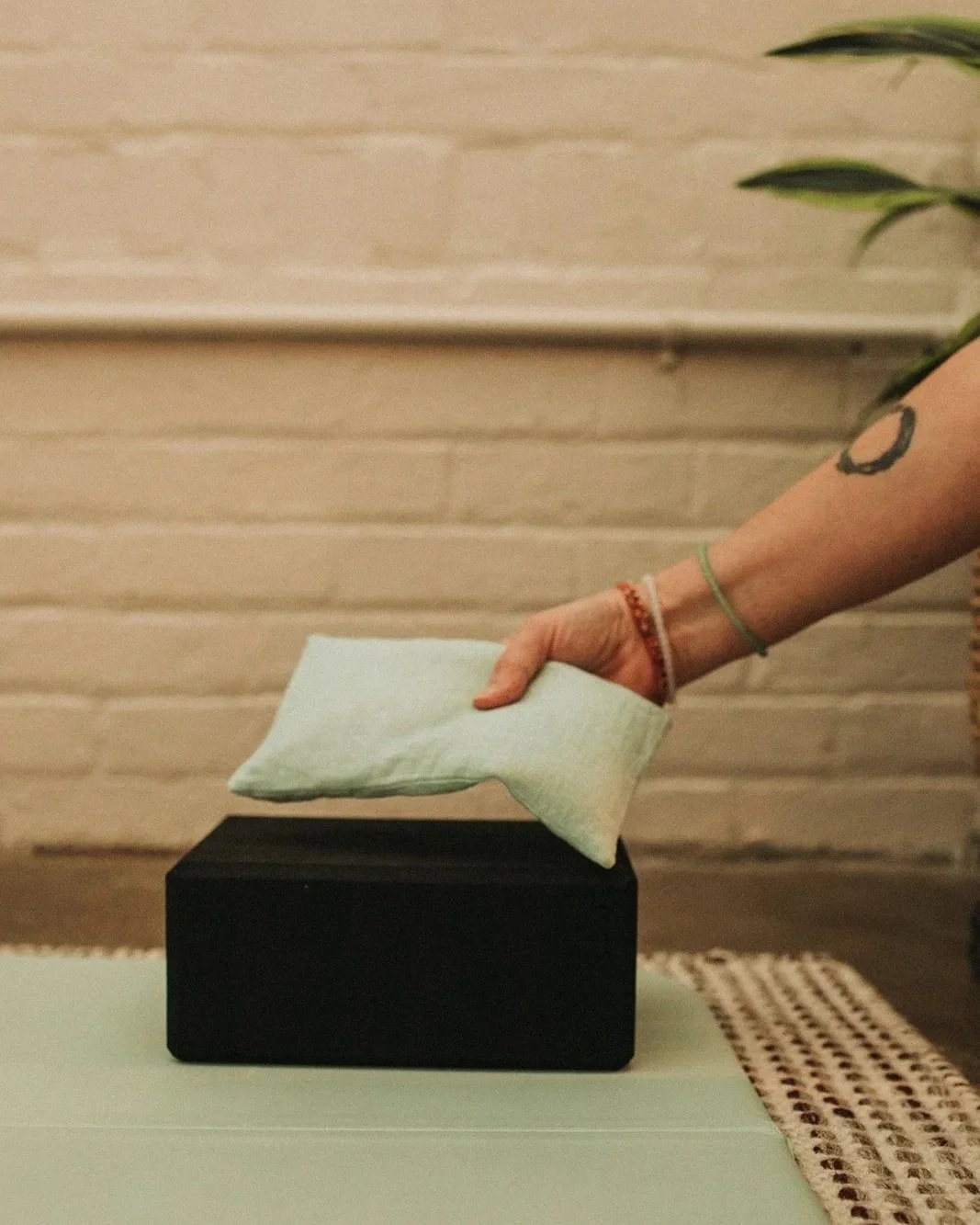 a yoga techer with an enzo tattoo on her arm is placing a green eye pillow on a black yoga block.