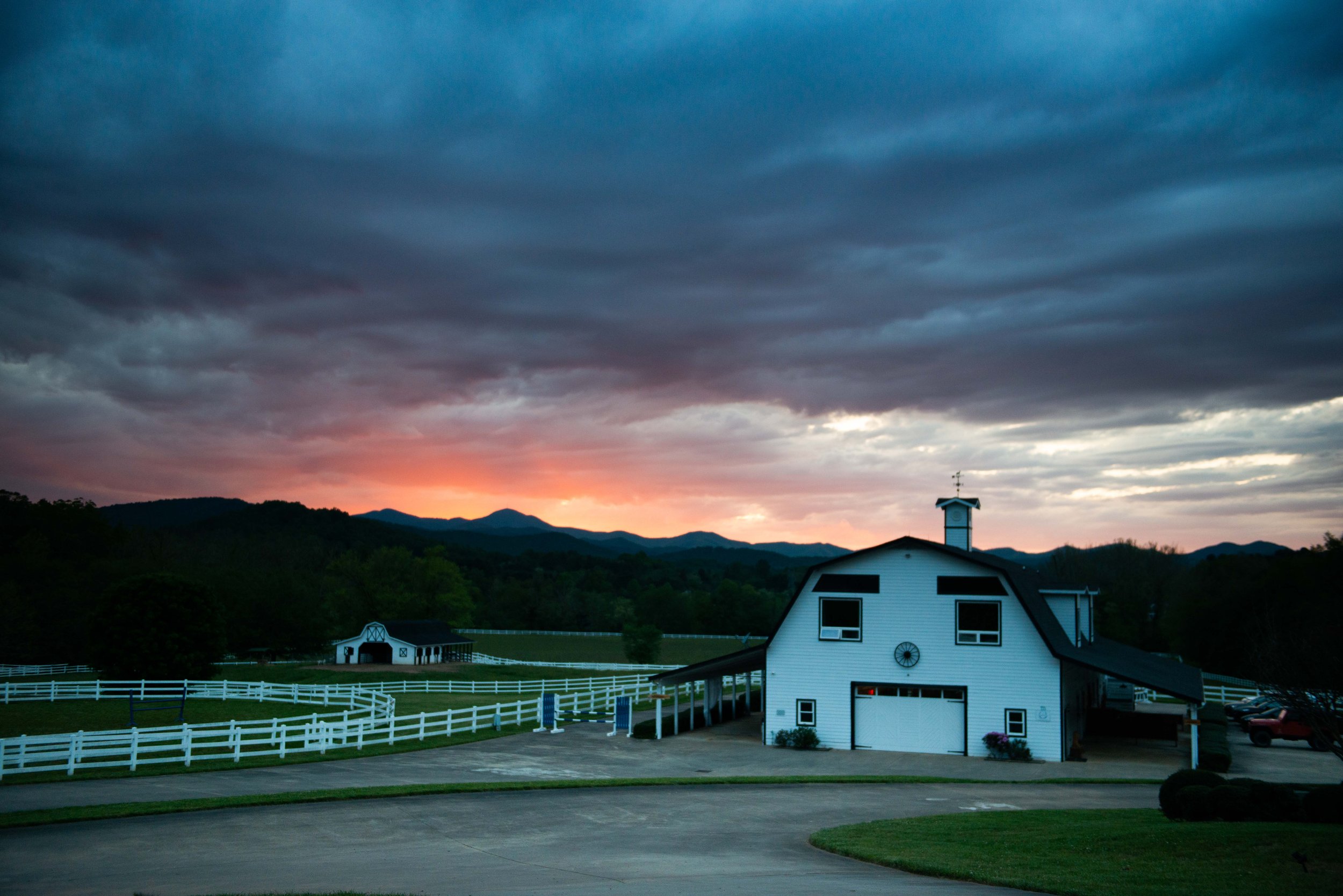 A white barn with black trim and a cupola, surrounded by white fencing, with mountains in the background and a dark, cloudy sky at sunset.