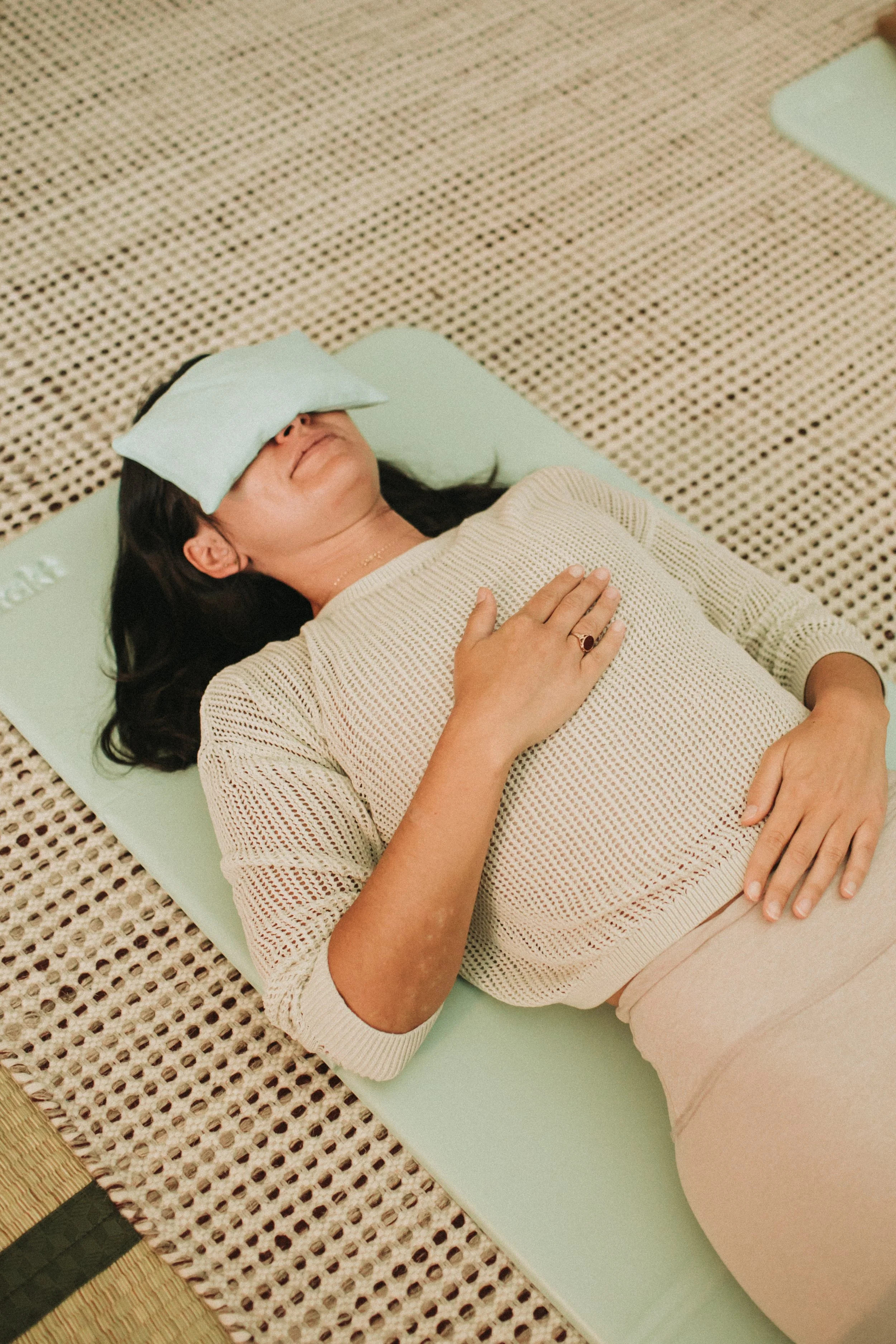 young wopmena with dark hair laying on a yoga mat with an eye pillow and hand over her heart