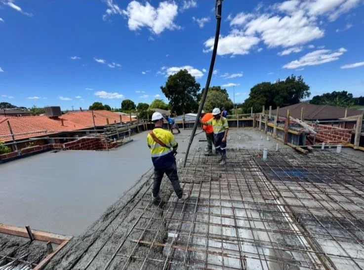 Construction workers on a building site pouring and leveling concrete over a steel rebar framework under a blue sky with scattered clouds.