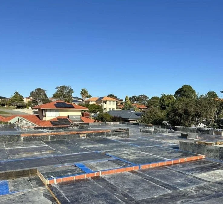 View of a rooftop under construction, with roofing materials and tools, overlooking a neighborhood with houses and trees under a clear blue sky.