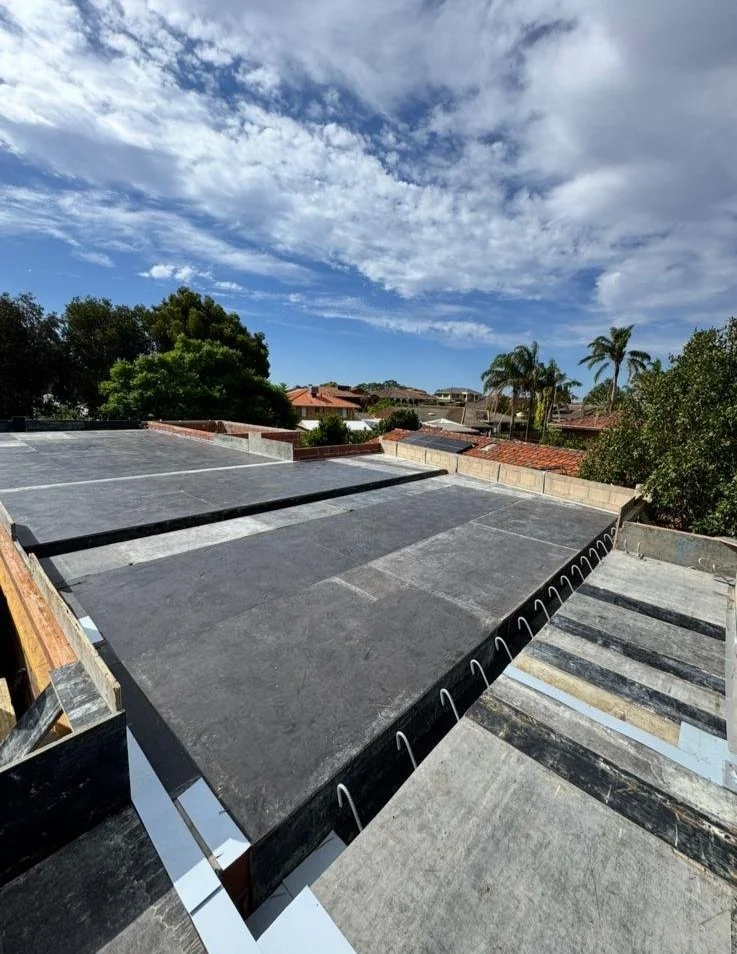 View of roofs under construction with black sheathing, orange roofing tiles visible on some neighboring buildings, palm trees, and a partly cloudy sky.